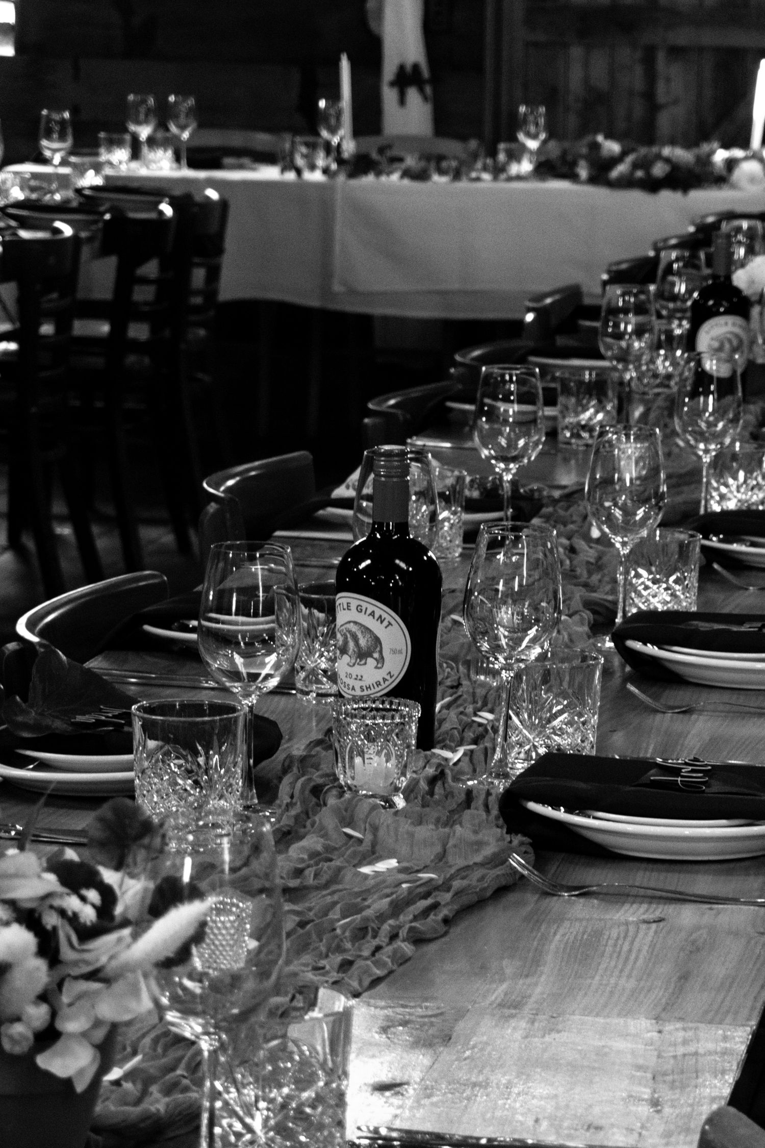 Black and white photo of an elegantly set dining table with crystal glasses, plates, and a wine bottle in a restaurant.