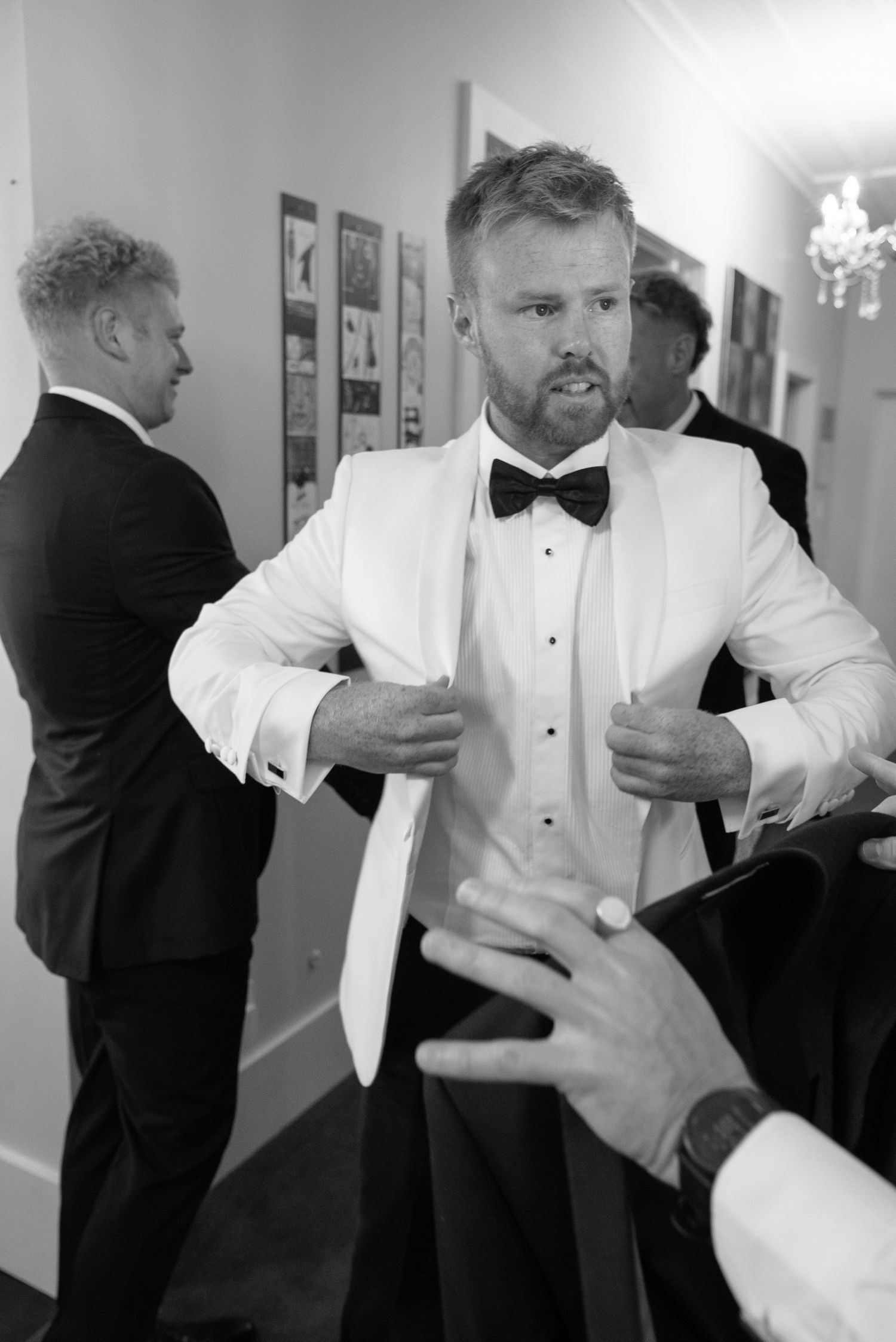 Black and white photo of a groom in white tuxedo jacket and bow tie adjusting his suit before wedding ceremony.
