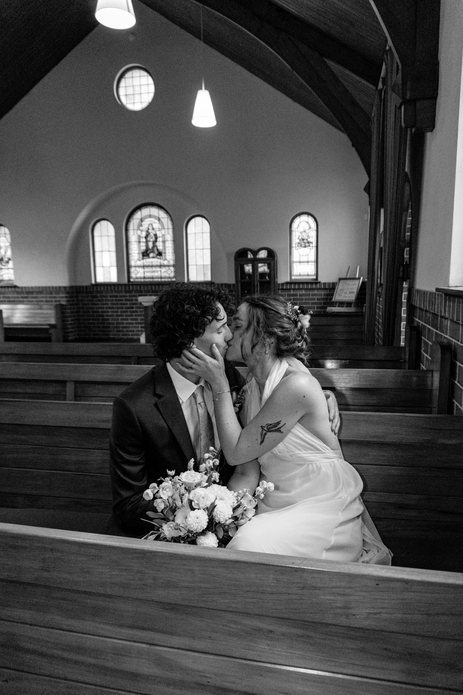 Black and white photo of a bride and groom sharing a kiss in a church pew, bride holding a bouquet.