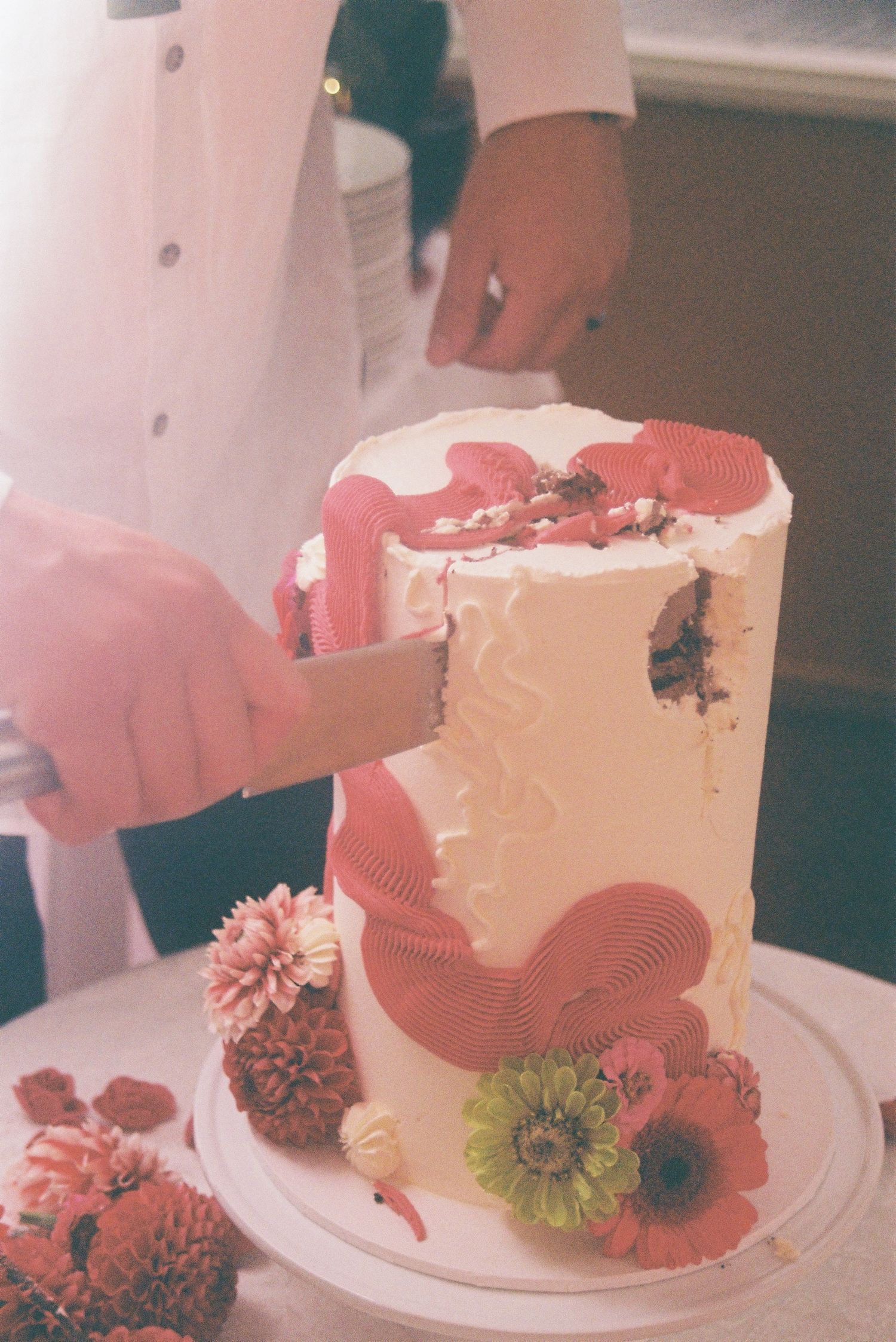 A chef cutting a tall white wedding cake decorated with pink drizzle and fresh flowers including dahlias and poppies.