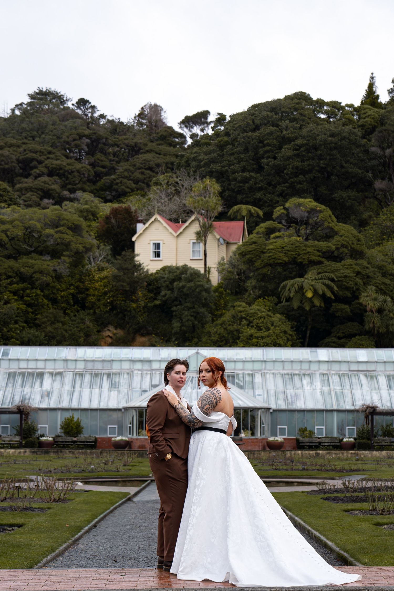 Two brides pose in a lush garden with a greenhouse and hillside mansion backdrop on their wedding day.