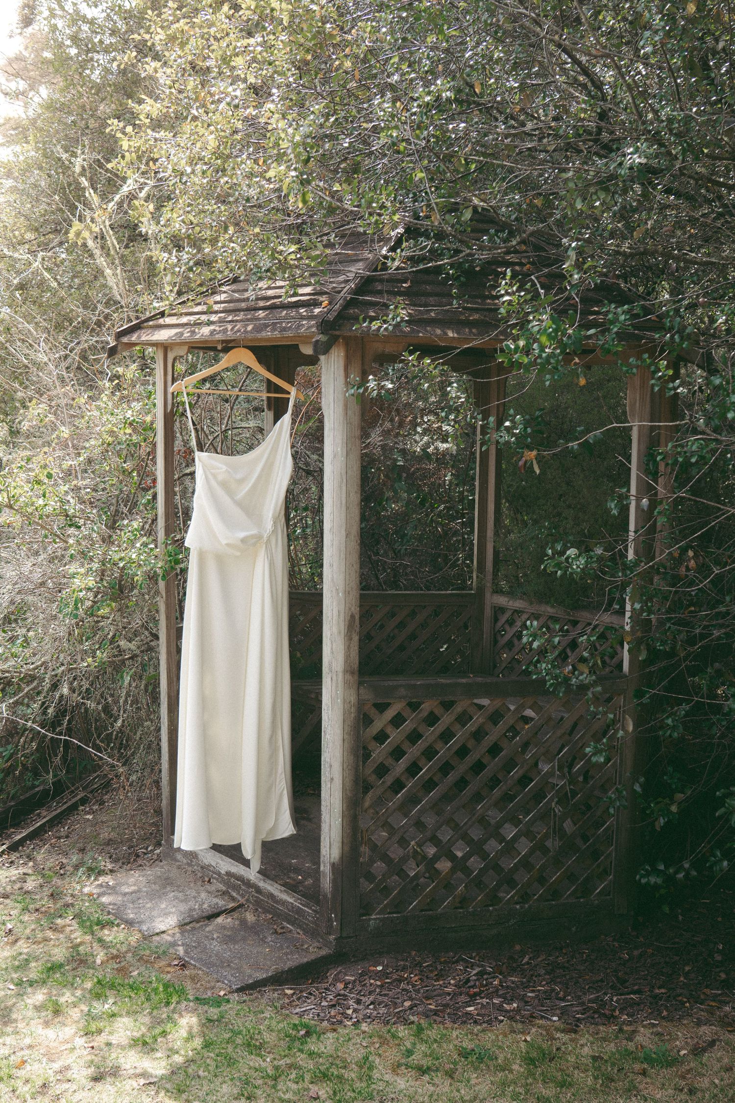 White wedding dress hanging on wooden hanger in rustic garden gazebo surrounded by lush green trees.