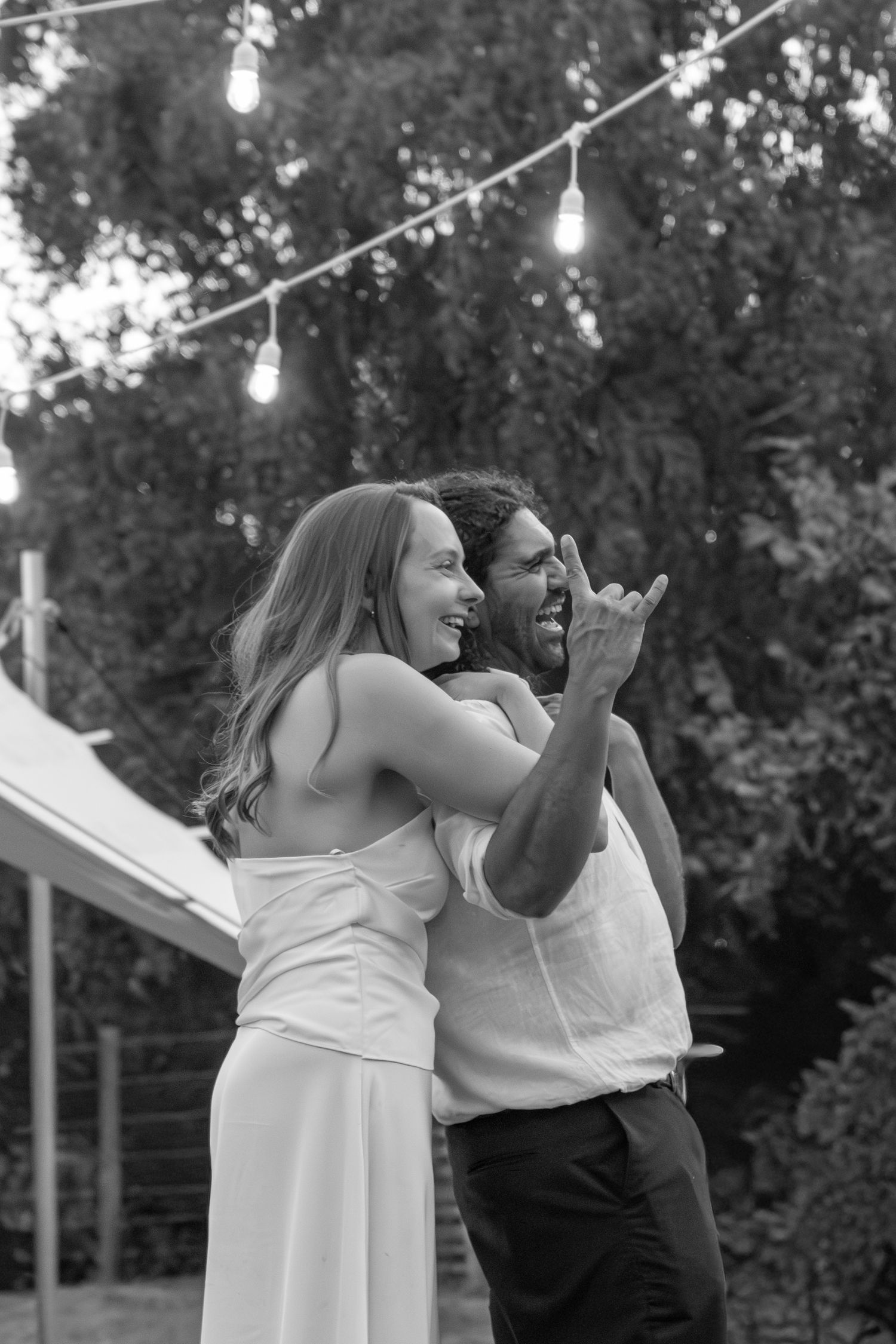 Black and white photo of a joyful couple dancing outdoors at a garden party with string lights.