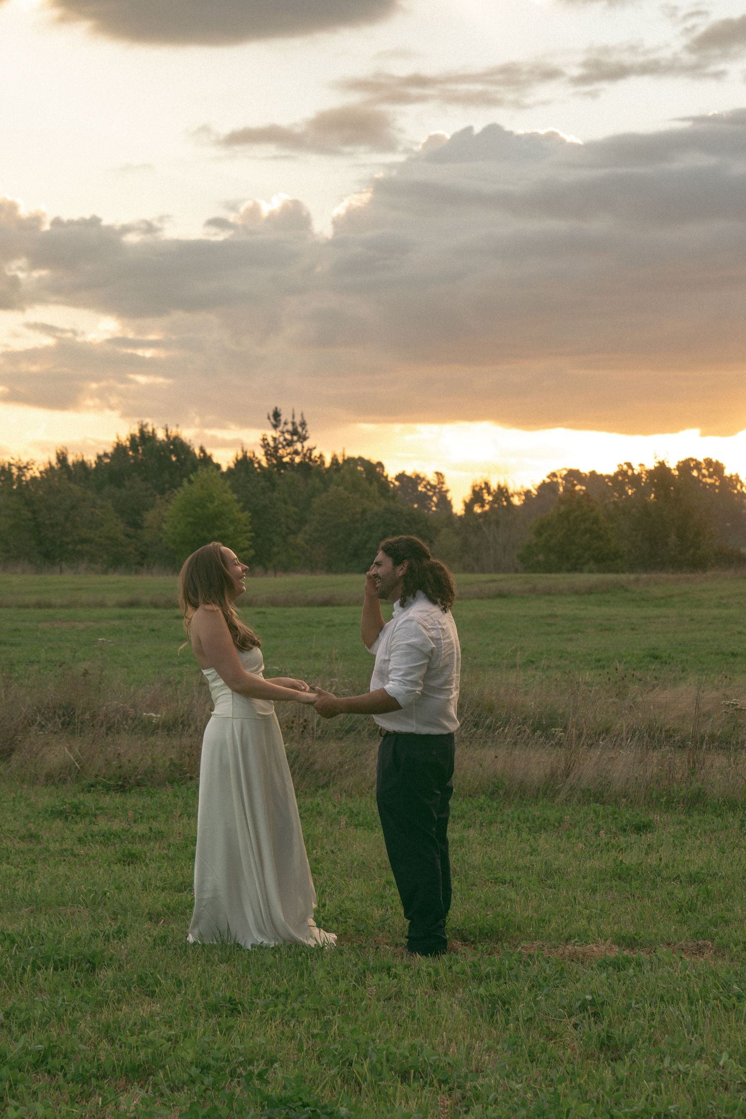 Bride and groom hold hands in a green field at golden sunset, sharing a romantic outdoor wedding moment.