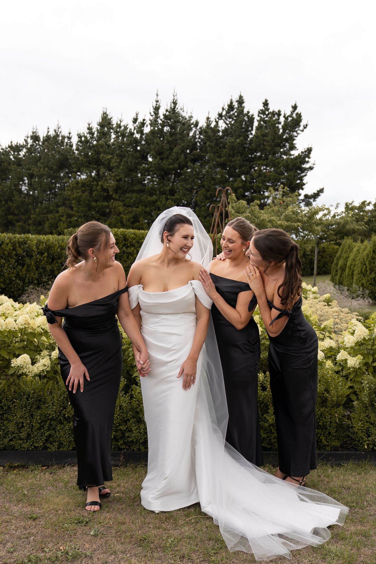 Bride in white gown and veil laughing with three bridesmaids in black dresses in a lush garden setting.