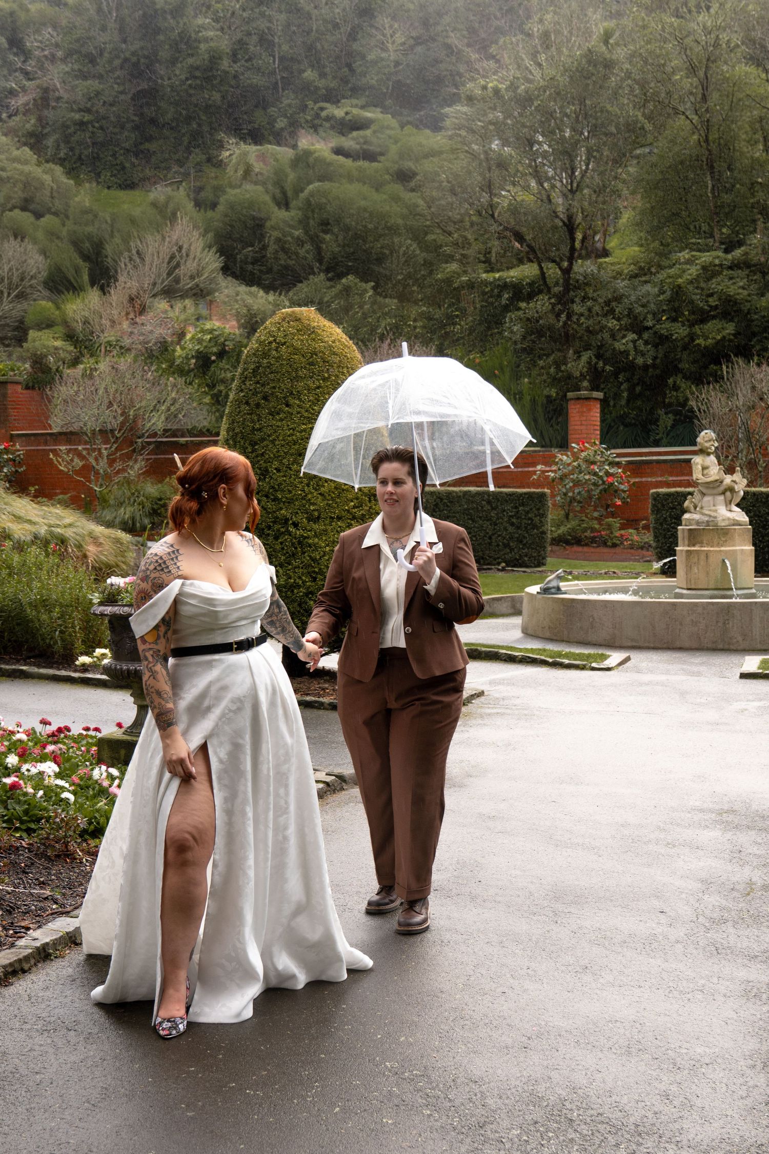 Two brides walk in a garden, one in a white gown and one in a brown suit holding a clear umbrella.