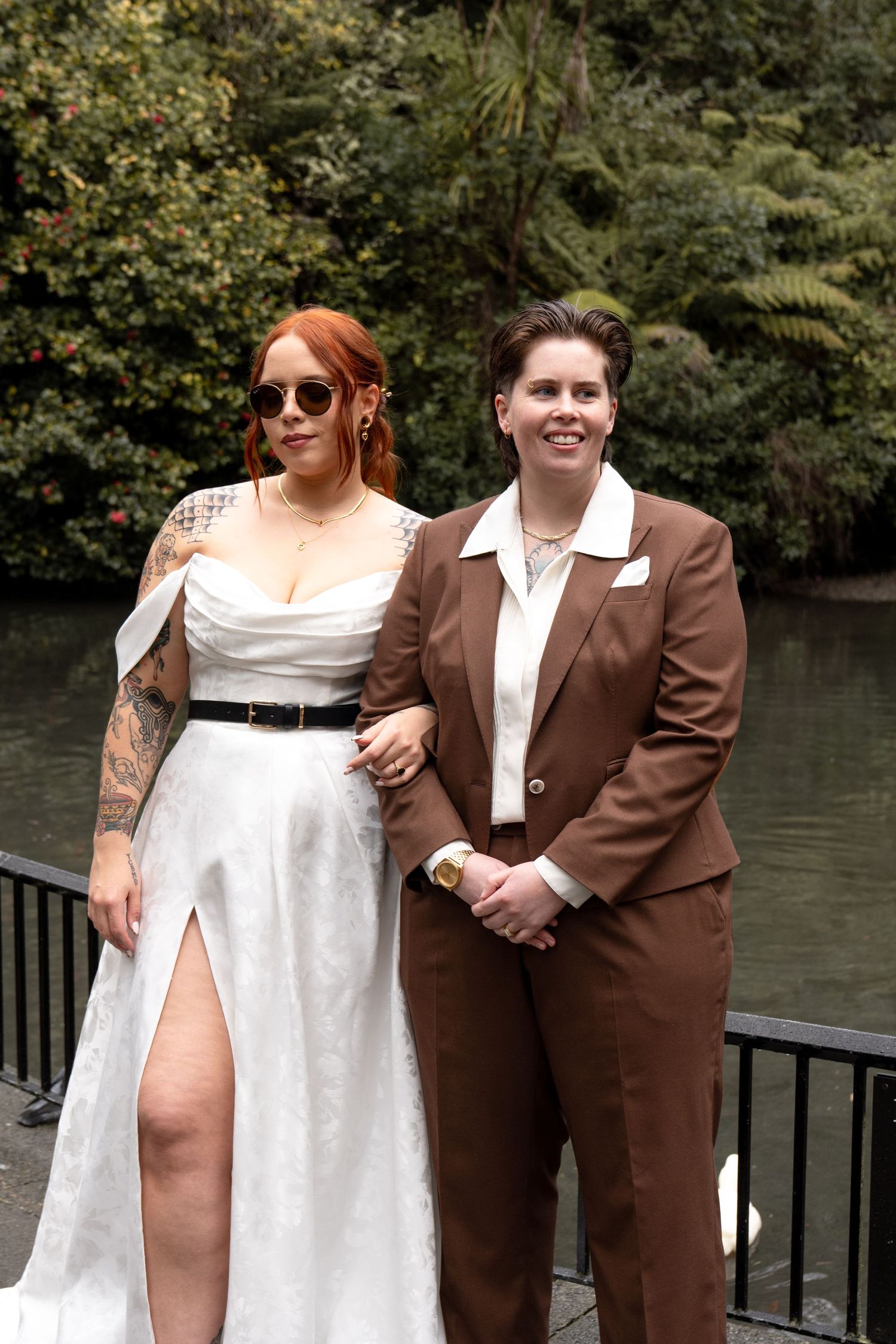 Two brides pose by a pond, one in a white gown with tattoos, the other in a brown suit, surrounded by lush greenery.