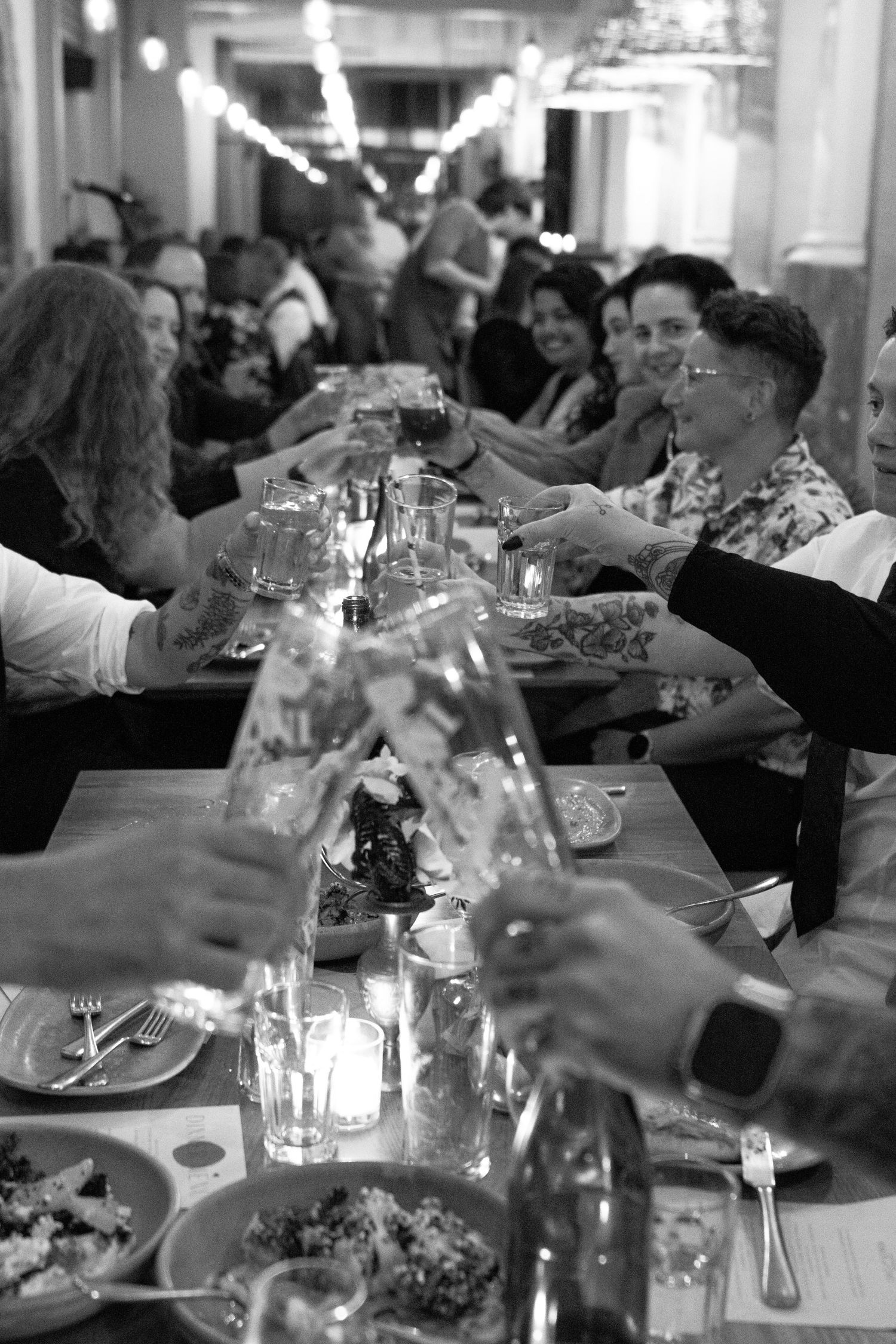 Black and white photo of guests toasting at a long dinner table during a festive group dining event.