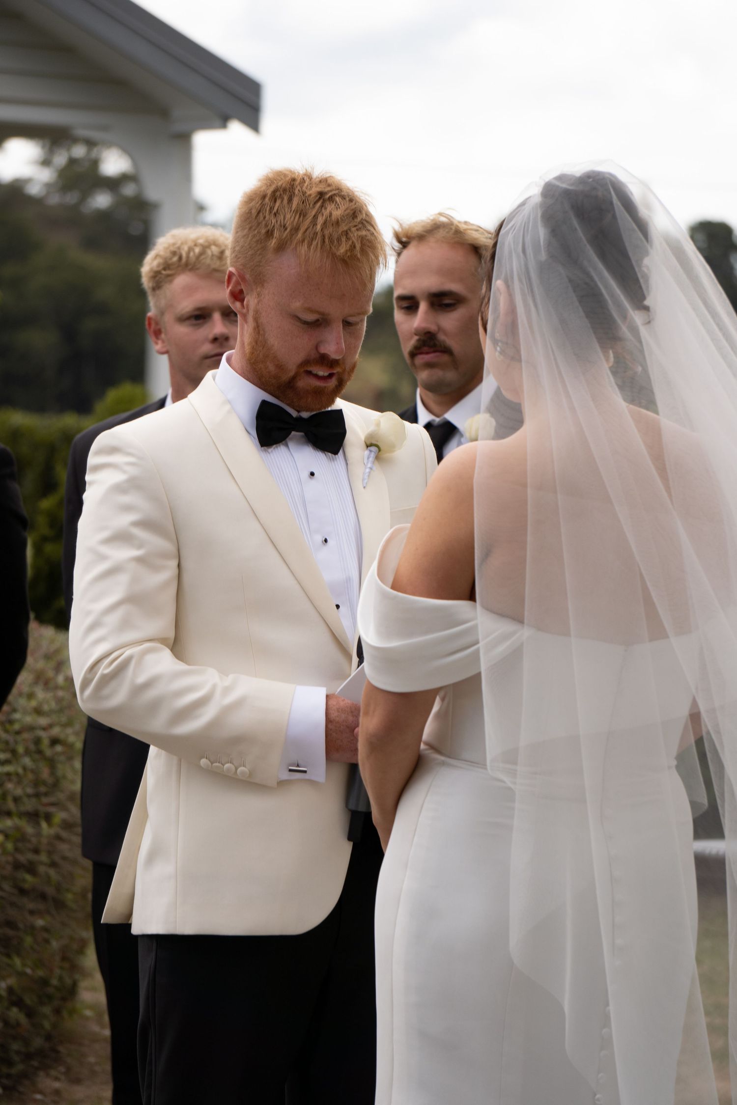 Groom in ivory tuxedo exchanges vows with bride in white gown and veil at outdoor wedding ceremony.