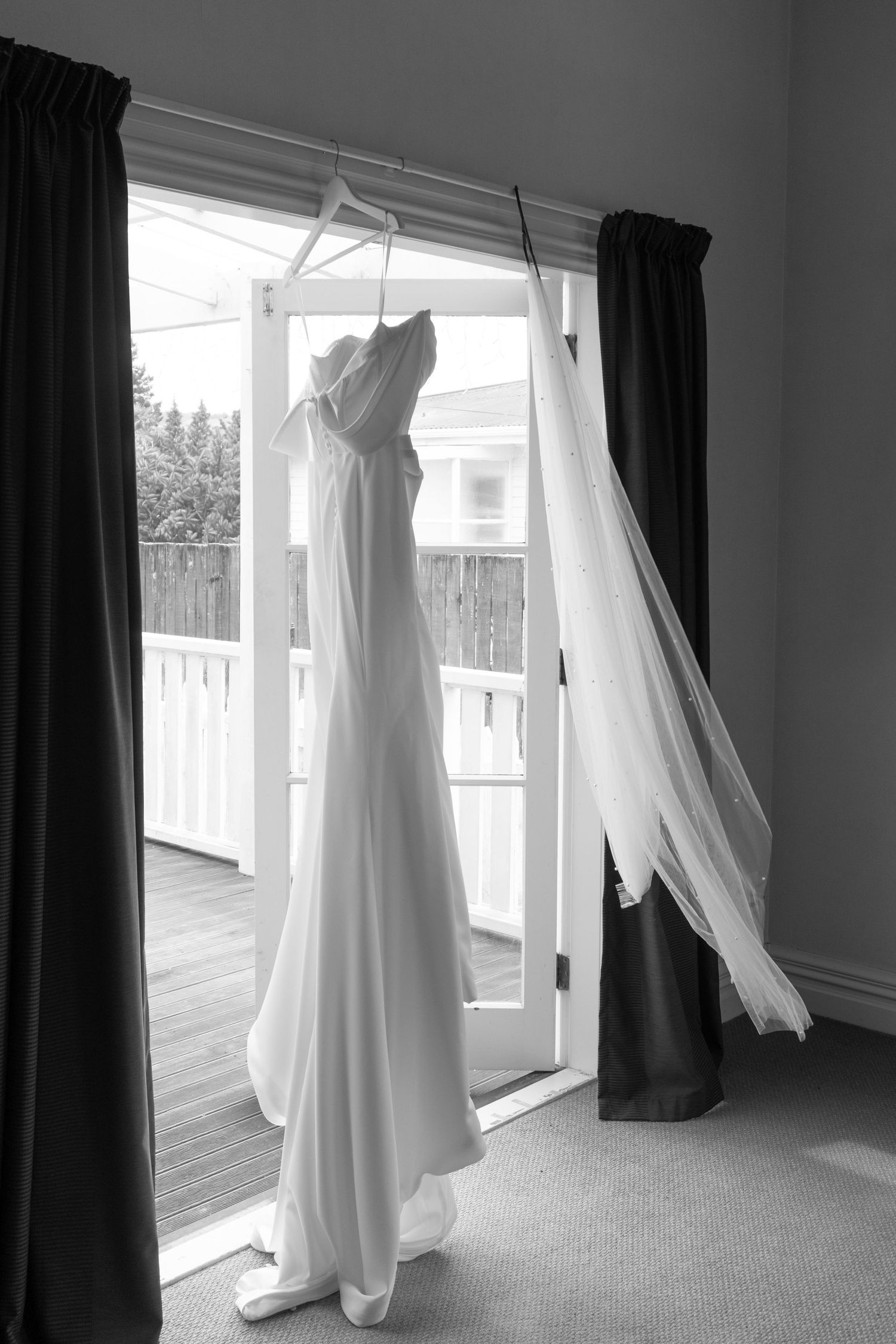 Black and white photo of an elegant wedding dress hanging on a door frame with sheer veil flowing beside it.