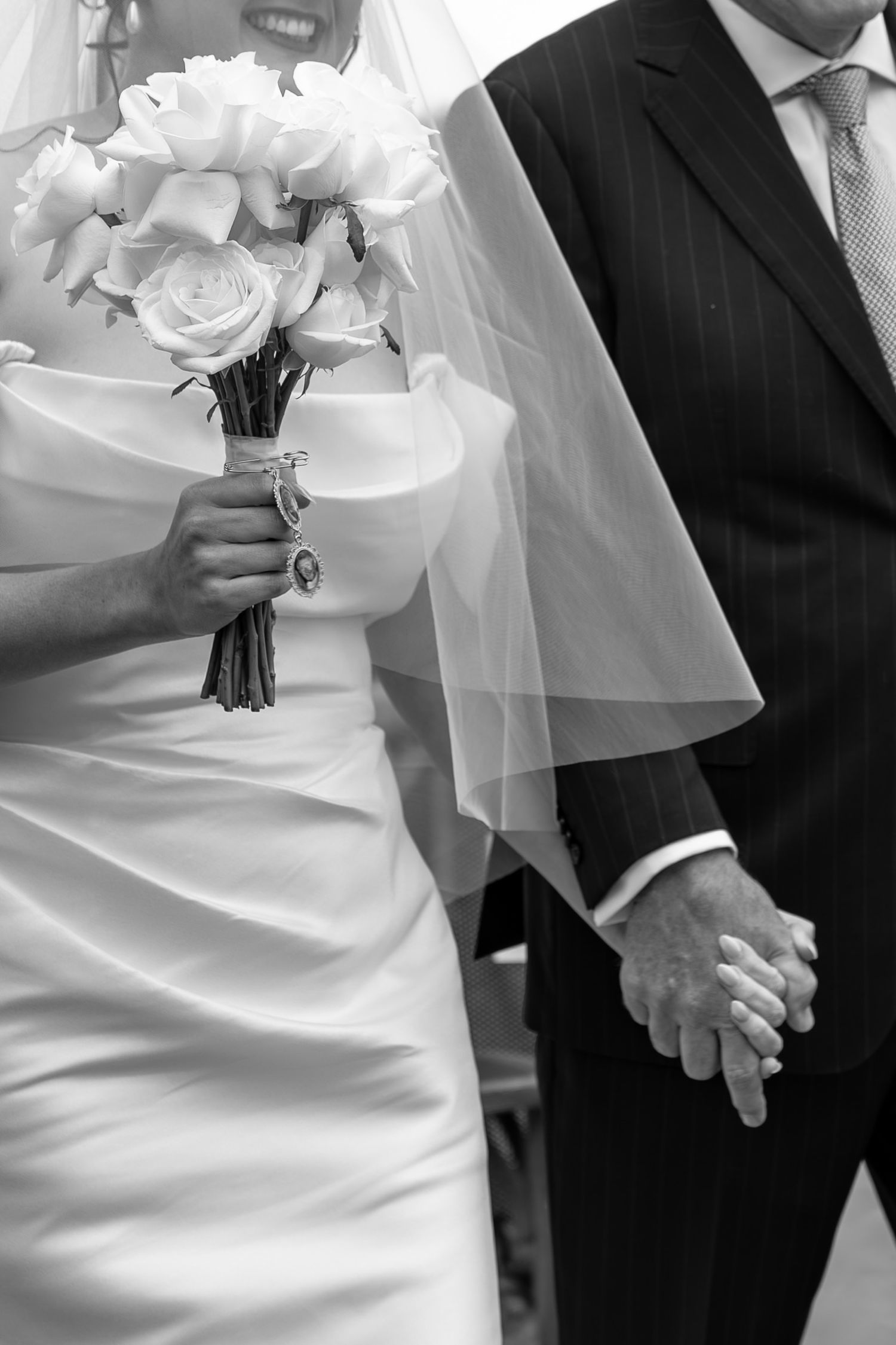 Black and white photo of bride holding rose bouquet and groom holding hands in wedding attire.