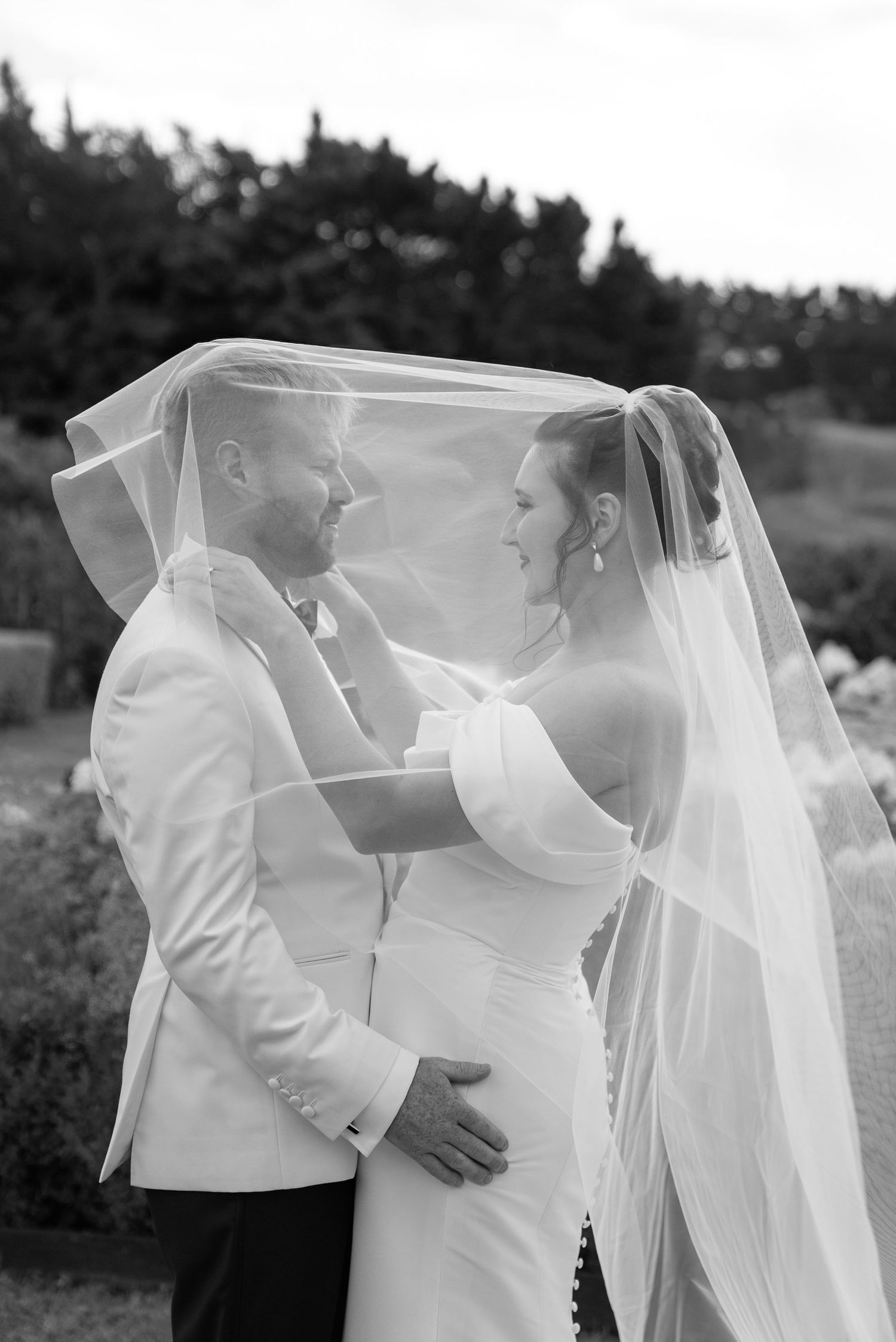 Black and white photo of bride and groom sharing a romantic moment under a flowing veil outdoors.
