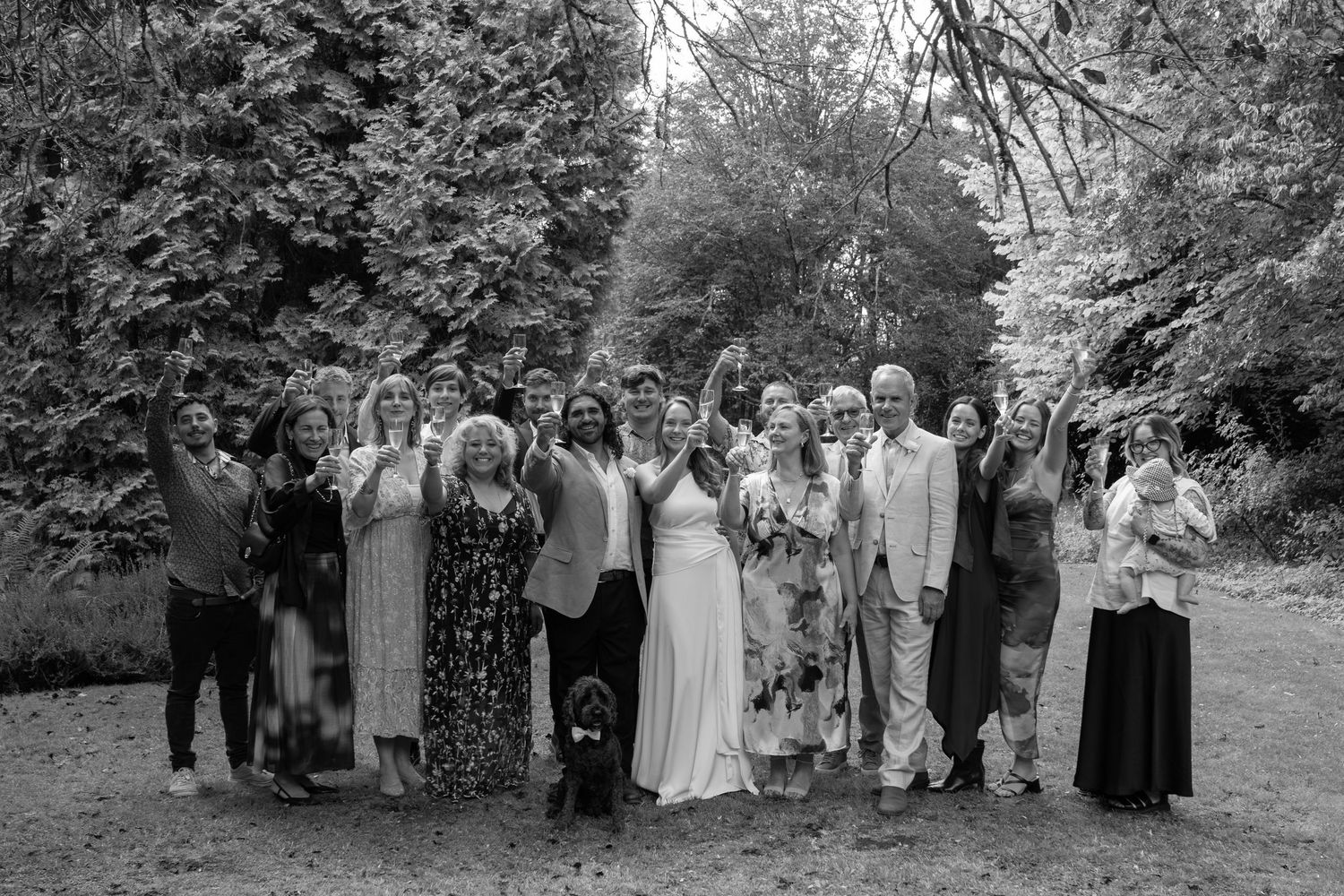 Black and white wedding group photo with bride and groom surrounded by guests celebrating outdoors among lush trees.