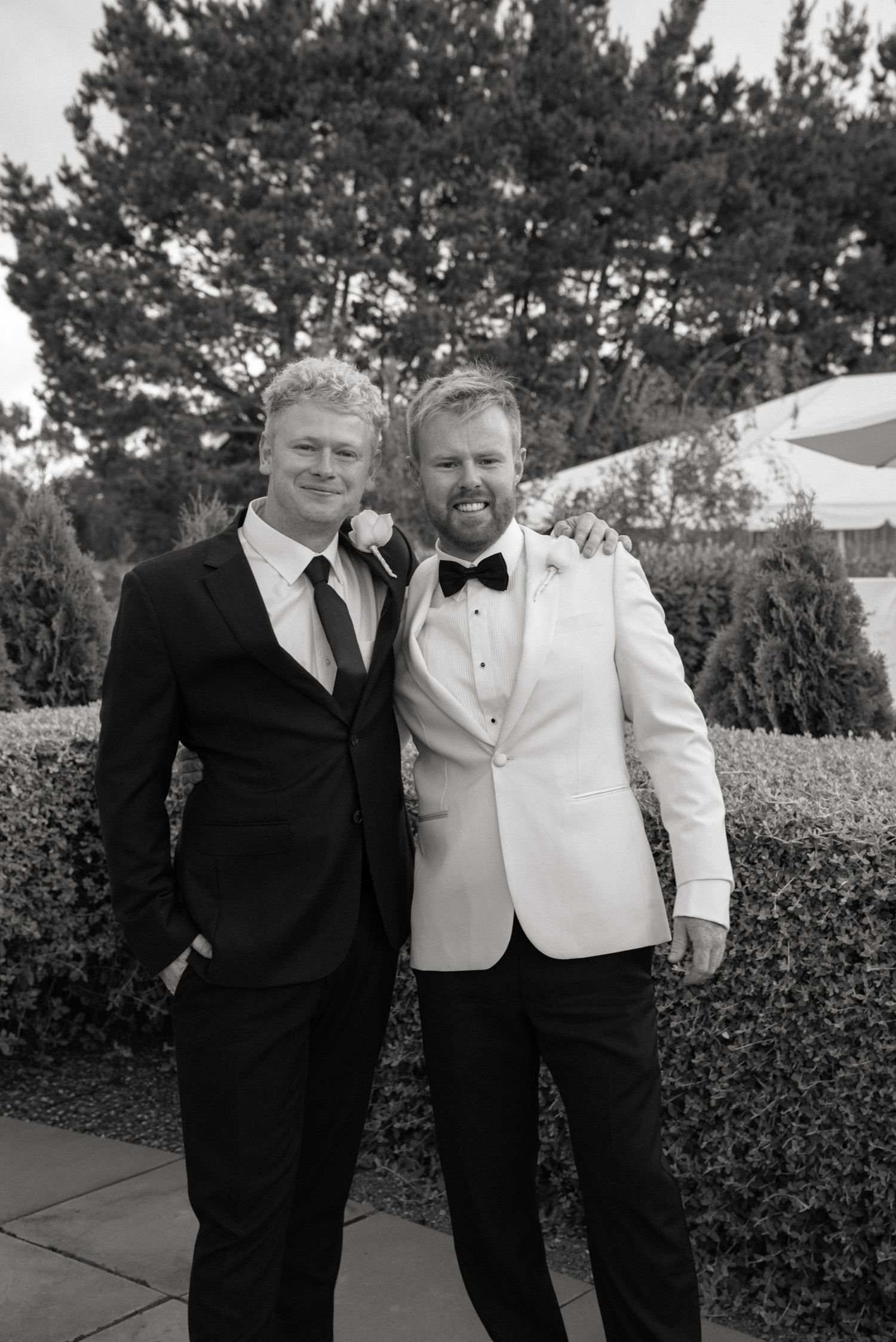 Two men in formal suits smile together outdoors at a wedding, one in black tuxedo and one in white jacket.