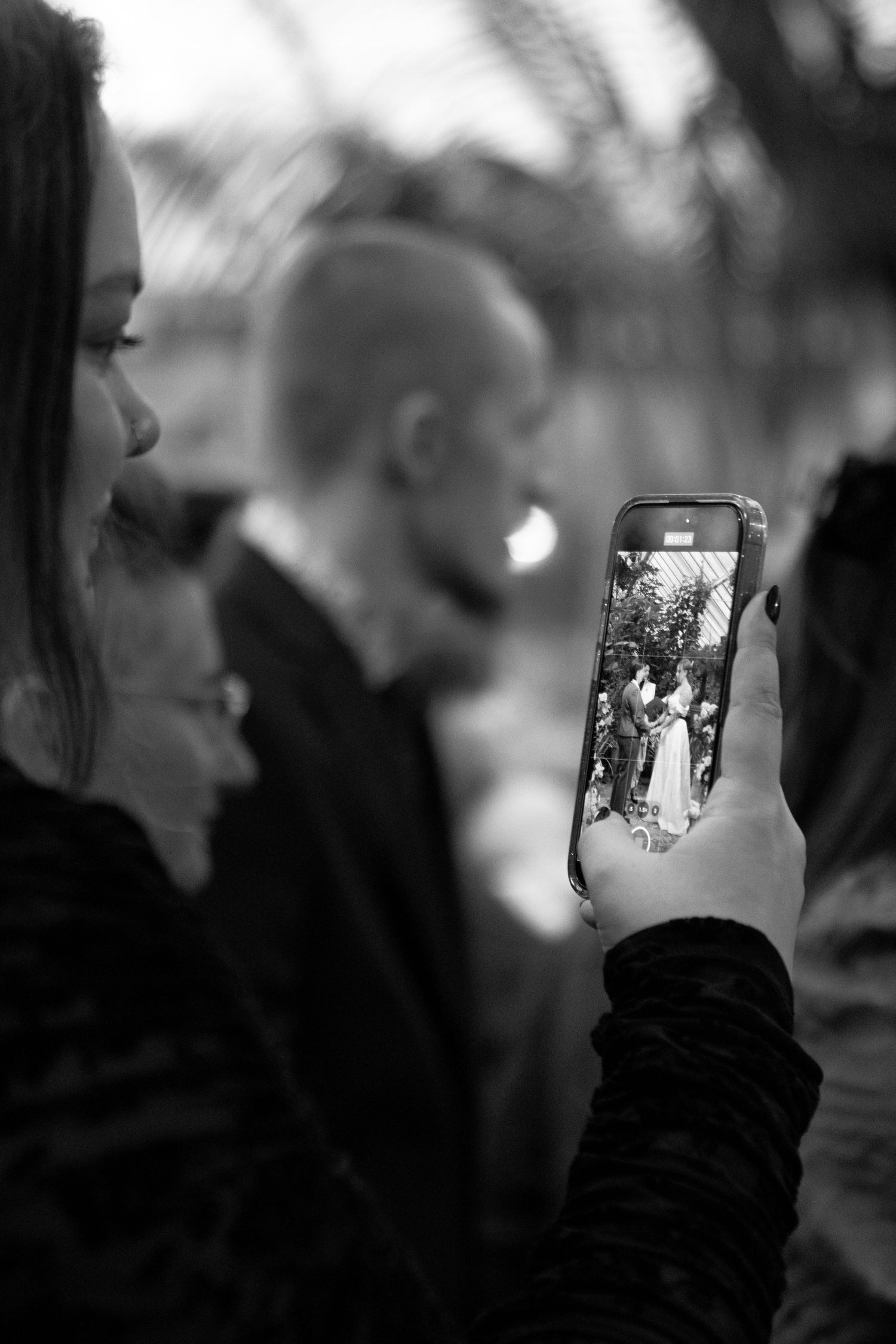 Black and white photo of a guest photographing a wedding ceremony on a smartphone at an outdoor event.
