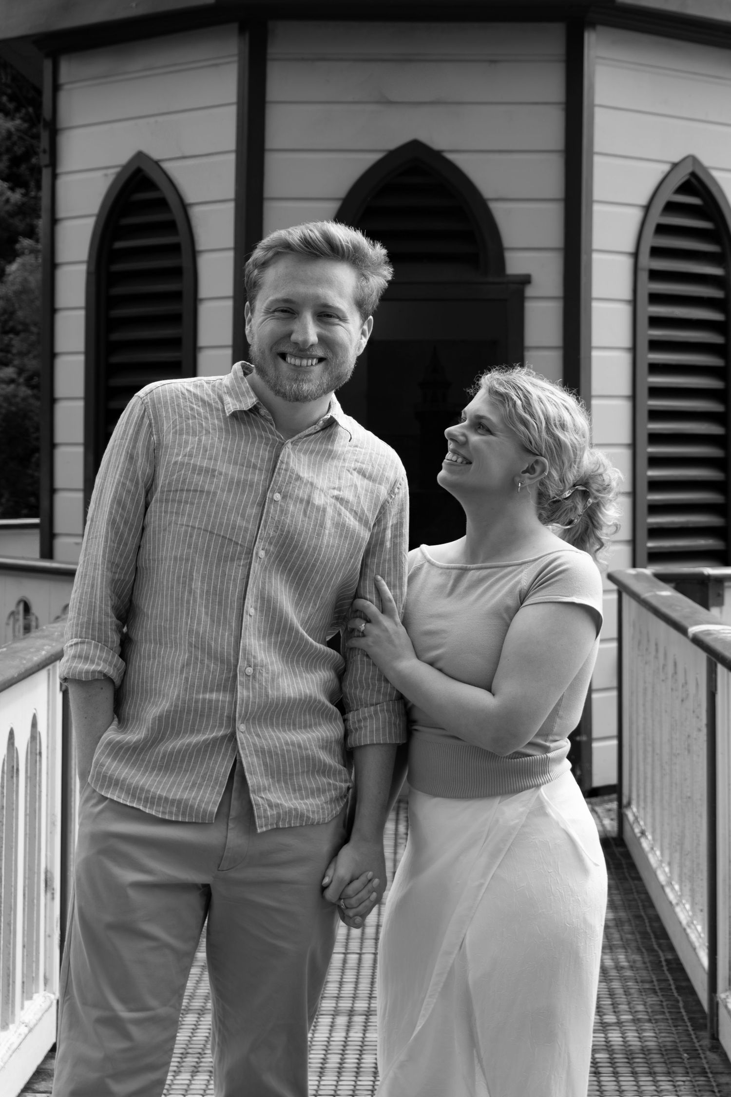 Black and white photo of a smiling couple holding hands outside a historic building with Gothic arch windows.