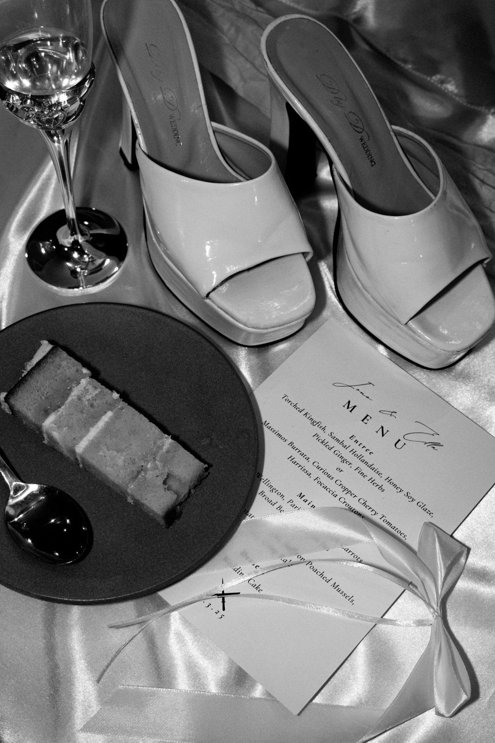 Black and white photo of elegant heels, wine glass, menu card, and dessert plate on satin fabric.
