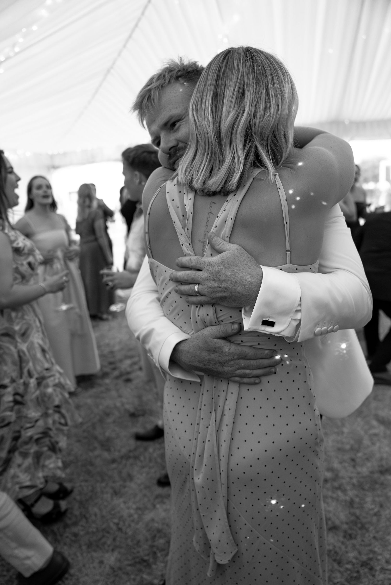 Black and white photo of a couple embracing warmly at an elegant outdoor wedding reception under a white tent.