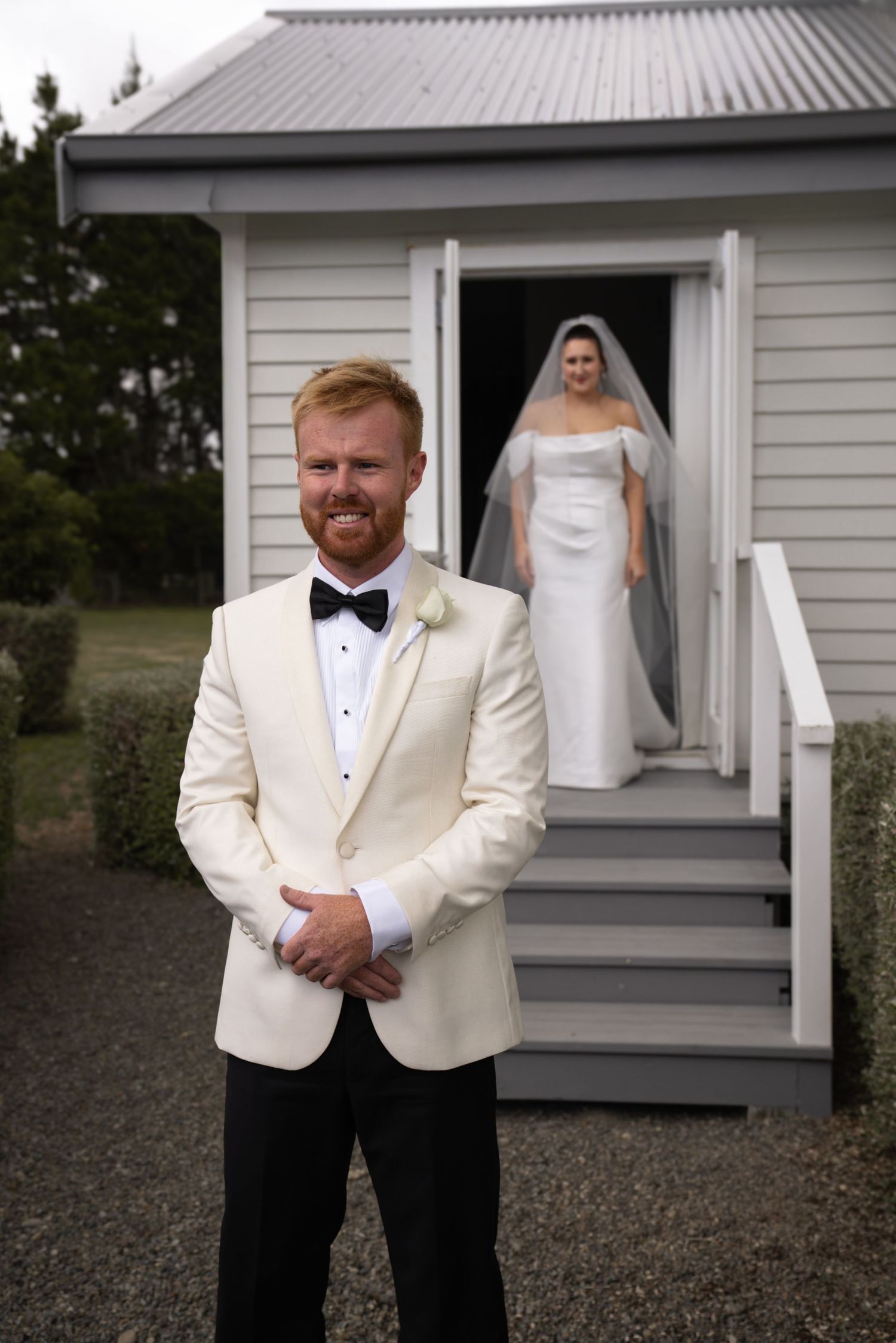 Groom in ivory tuxedo smiles outdoors as bride in white gown and veil emerges from chapel doorway behind him.