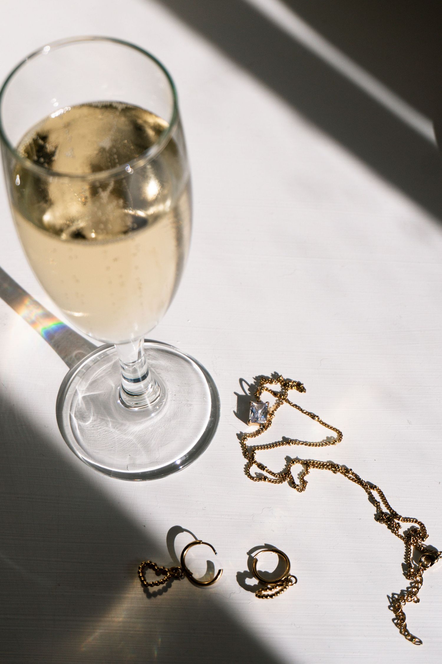 A glass of champagne beside gold jewelry including a necklace and earrings on a white surface.