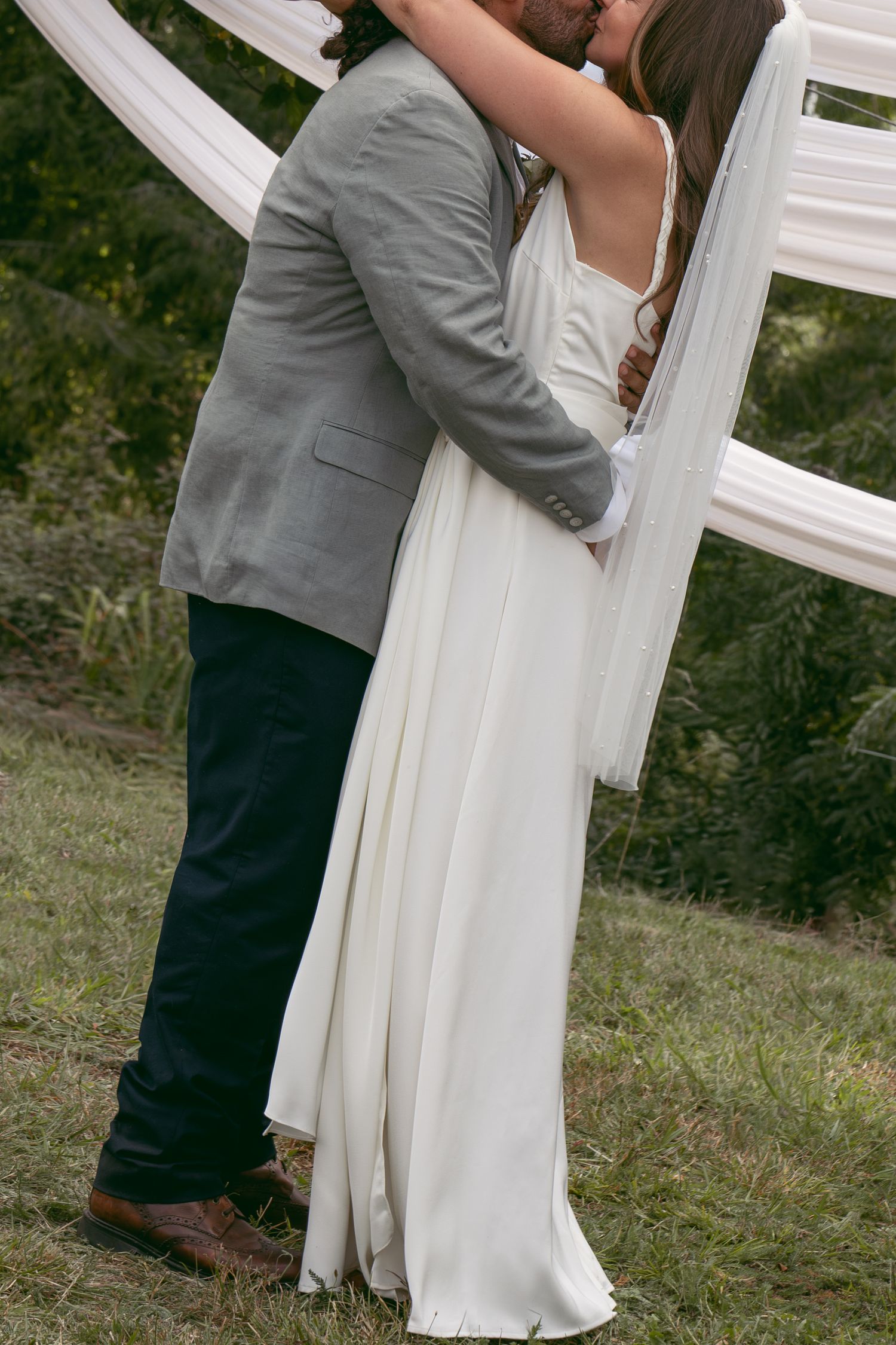 Bride and groom sharing a first kiss in white wedding dress and gray suit at outdoor ceremony.