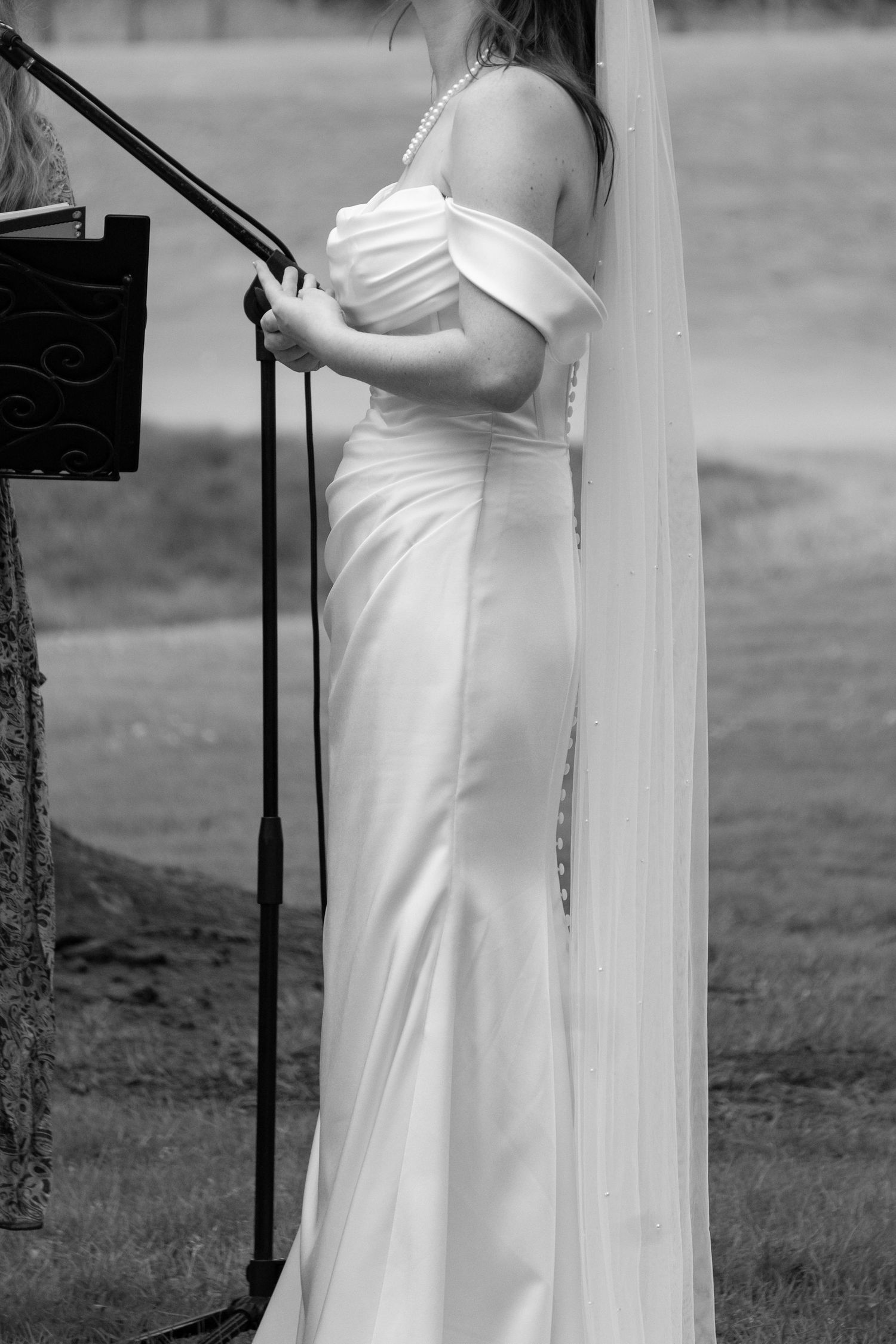Black and white photo of a bride in an off-shoulder white gown and veil standing at a microphone outdoors.