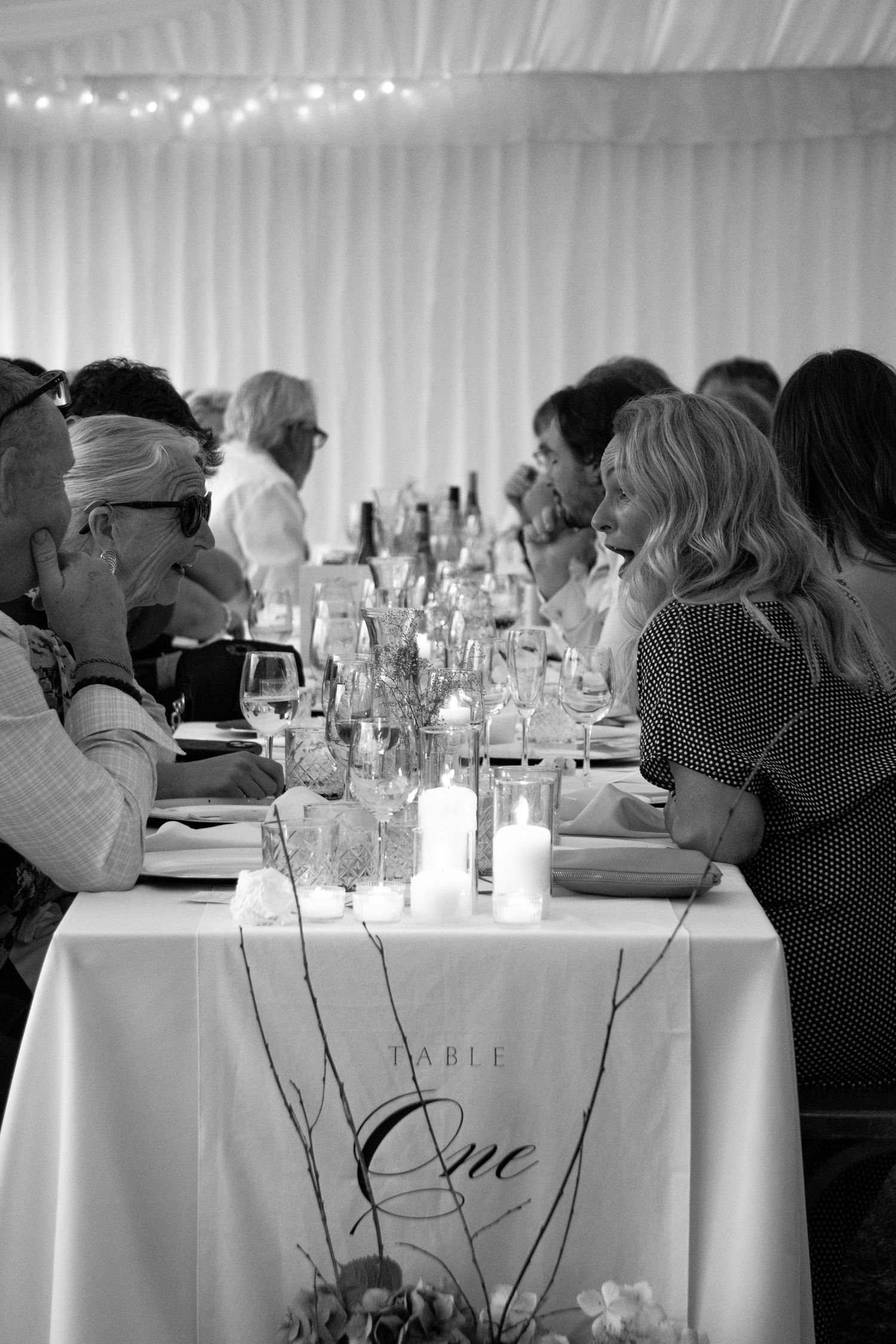 Black and white photo of wedding guests seated at Table One, adorned with candles and floral arrangements in a marquee.