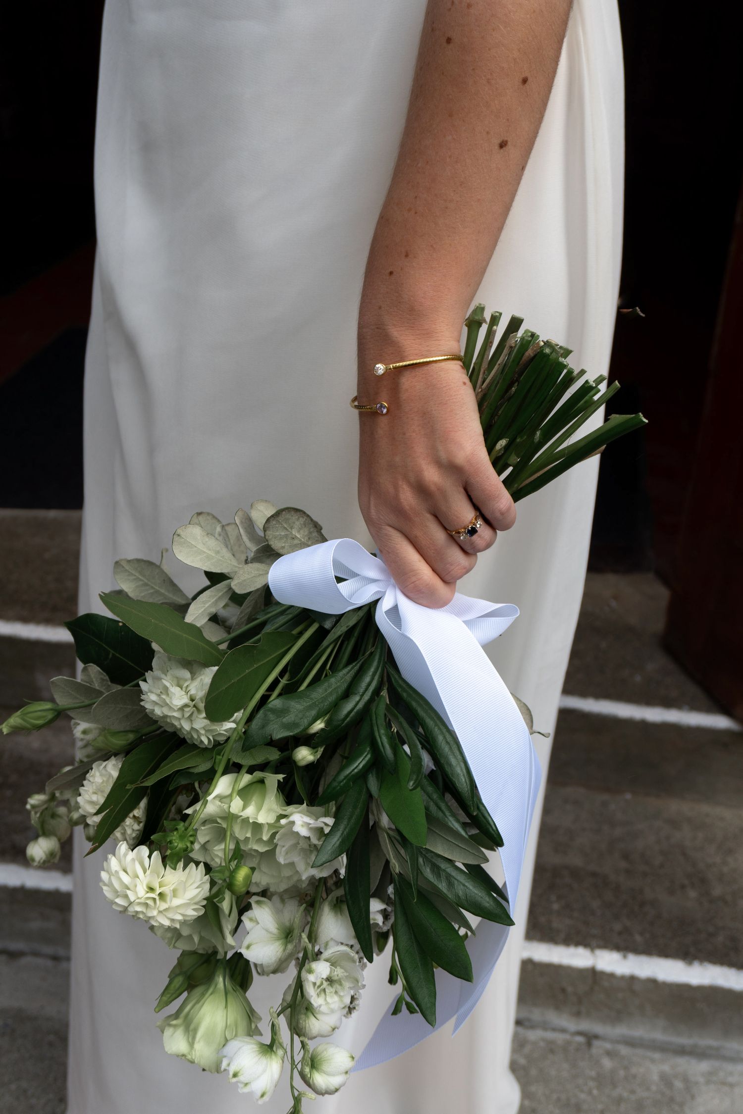 Bride in white dress holding elegant wedding bouquet of white flowers and green leaves tied with white ribbon.