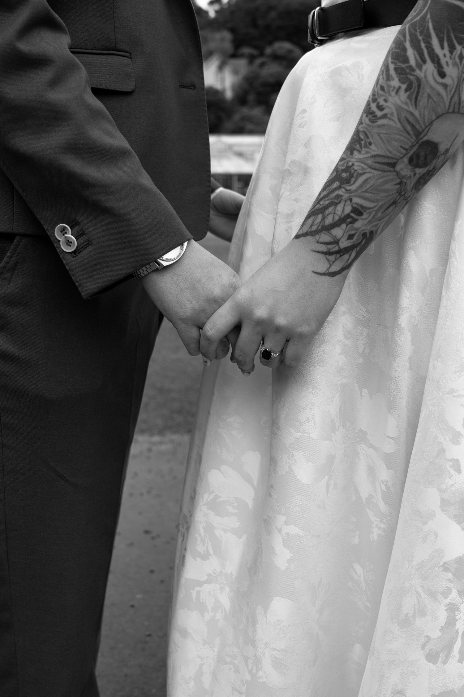 Black and white wedding photo of bride and groom holding hands, showing tattoo sleeve, ring, and formal attire.
