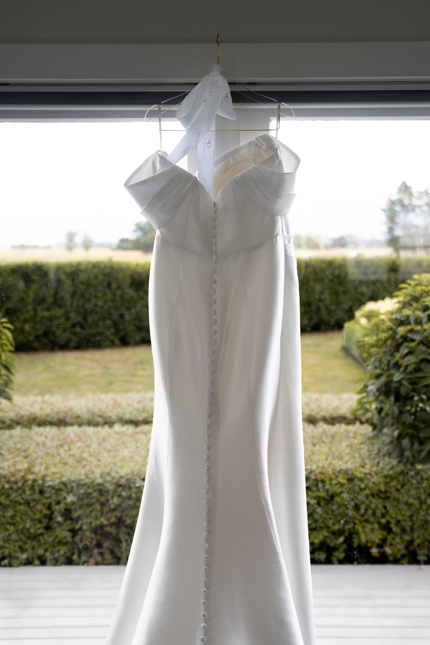 Elegant white strapless mermaid wedding dress hanging on a hanger in front of a window with garden views.