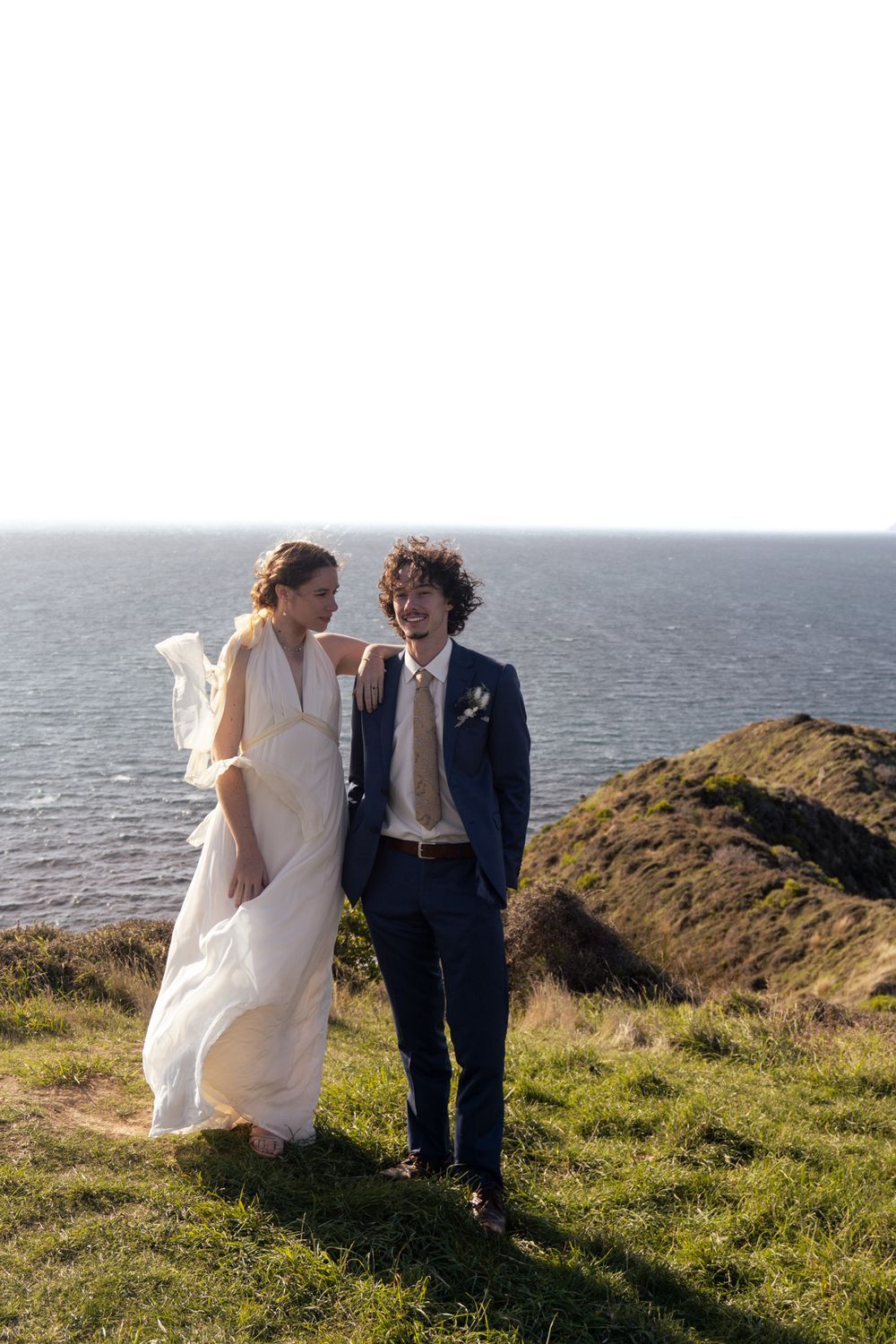 Wedding couple in formal attire stand on coastal cliff overlooking ocean.