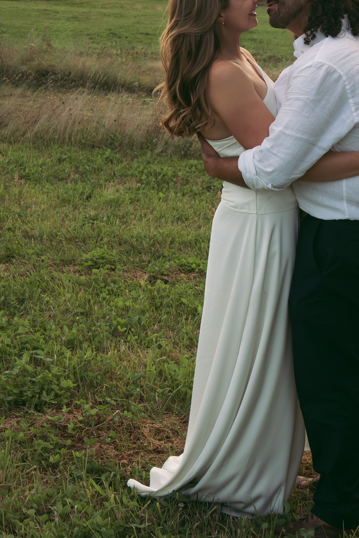 Couple embracing in a green field, bride in elegant white gown and groom in white shirt and dark pants.