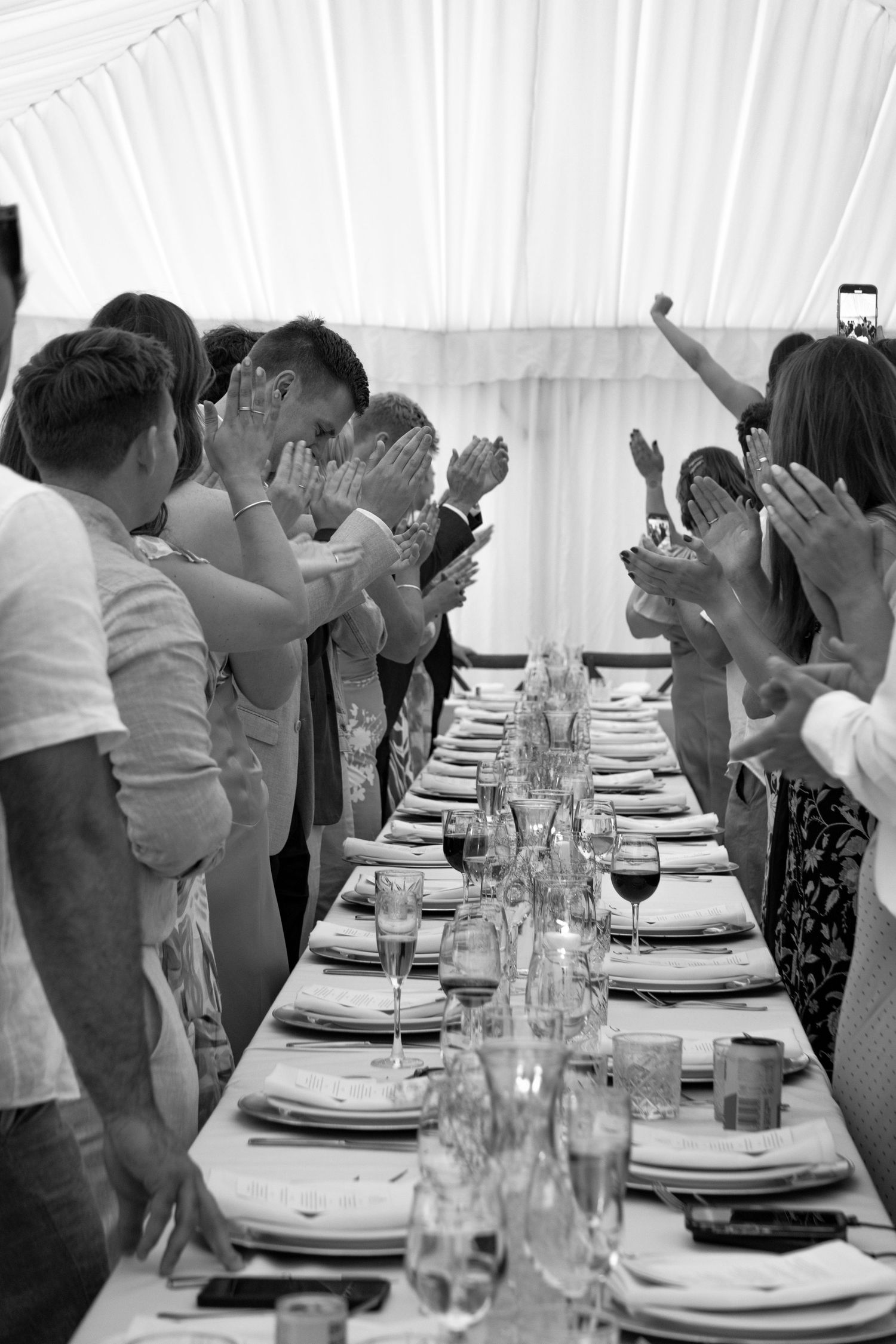 Black and white photo of guests clapping alongside a formal banquet table set with glasses and plates in a marquee.