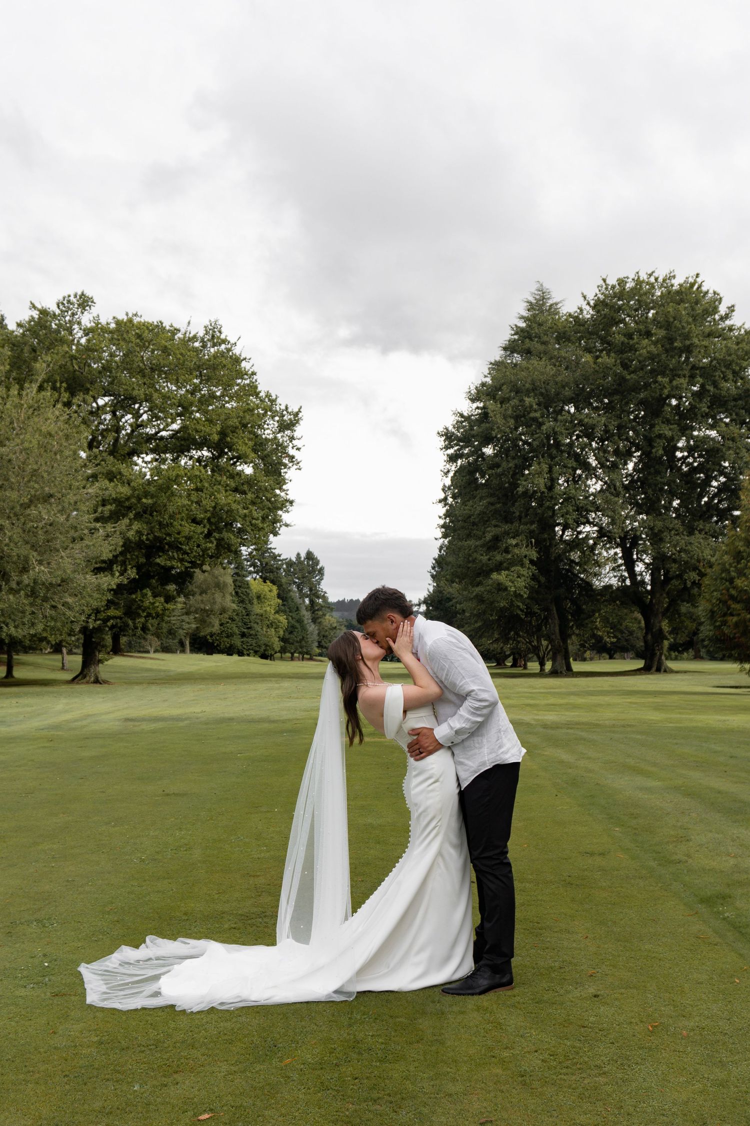Bride and groom share a romantic kiss on a lush green lawn, with the bride's long white veil trailing behind her.