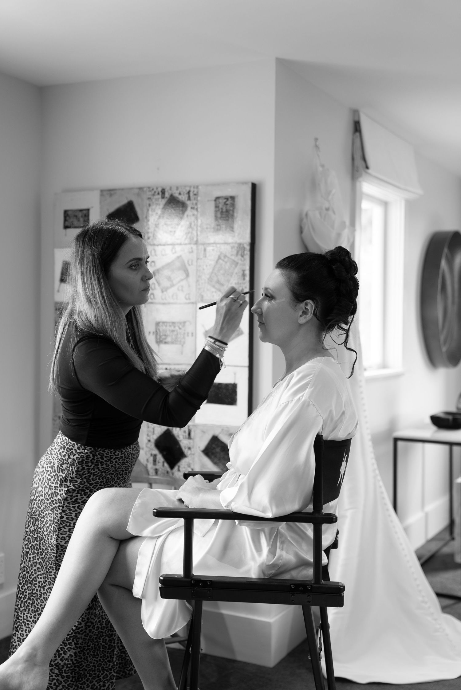 Makeup artist applying makeup to a bride in a white robe seated in a director chair during wedding preparations.