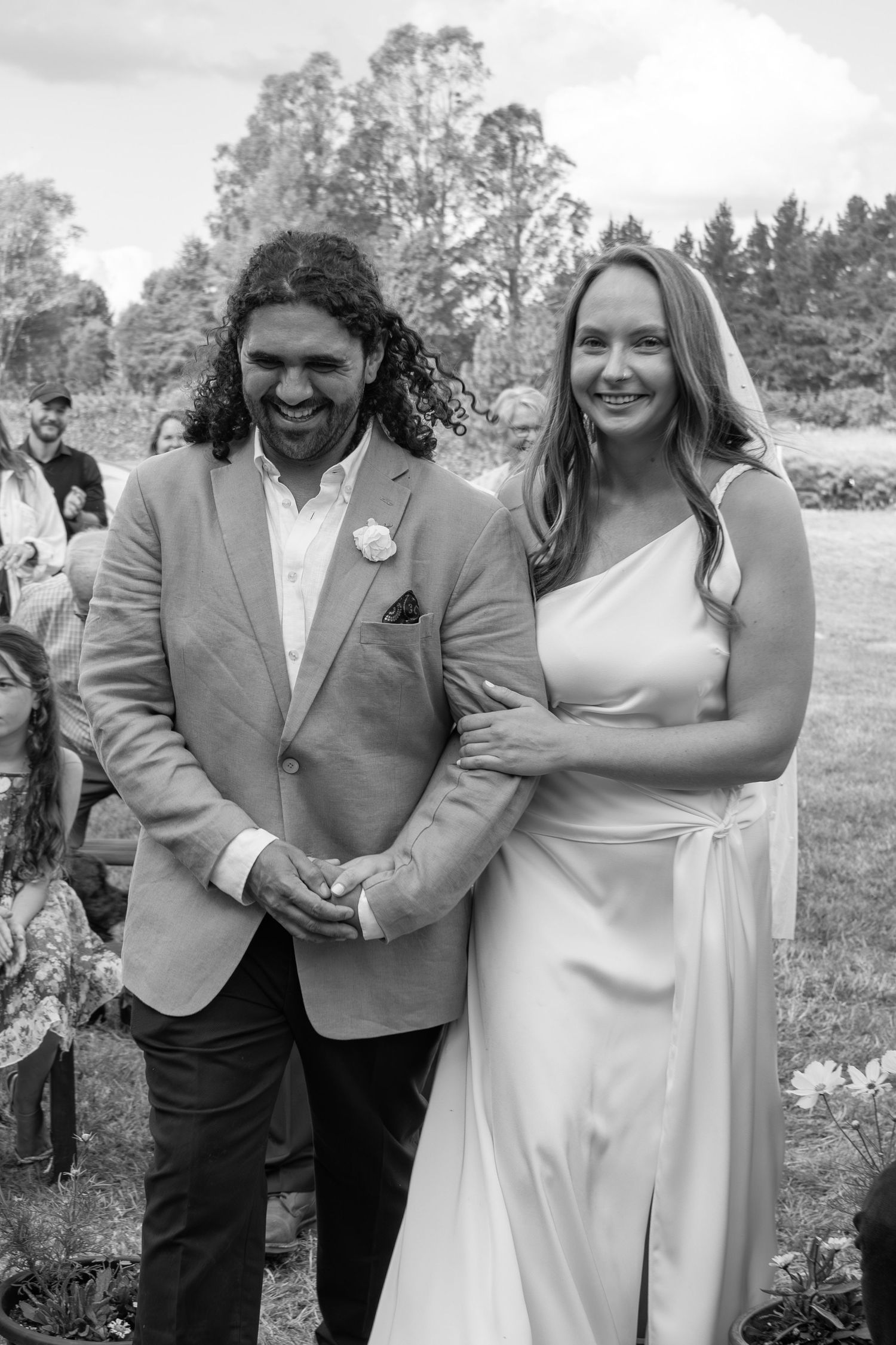 Black and white photo of a smiling bride in white gown and groom in suit holding hands at outdoor wedding ceremony.