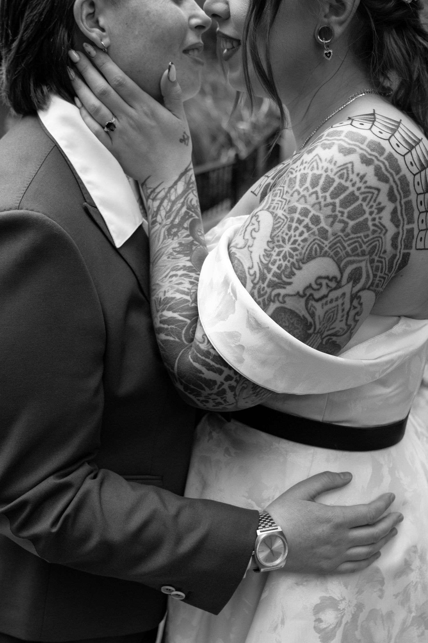 Black and white wedding photo of tattooed couple embracing, bride in white gown with floral sleeve tattoos, groom in suit.