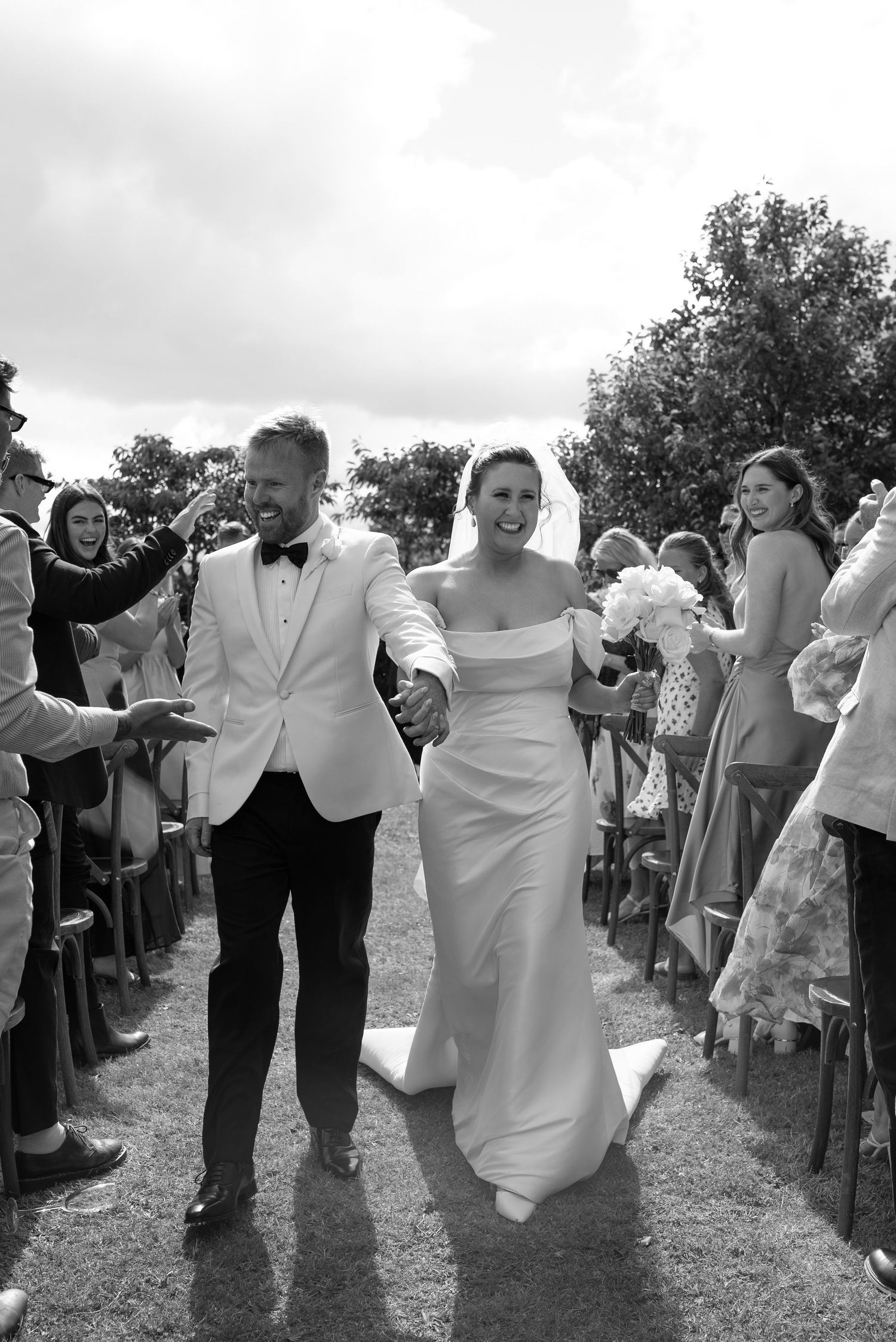 Black and white photo of a joyful bride and groom walking down the aisle hand in hand at an outdoor wedding ceremony.