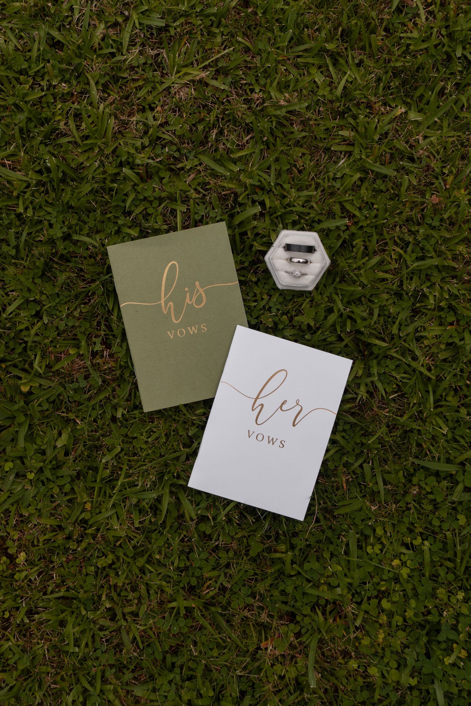 His and her vows cards with a hexagonal ring box displayed on lush green grass for a wedding ceremony.