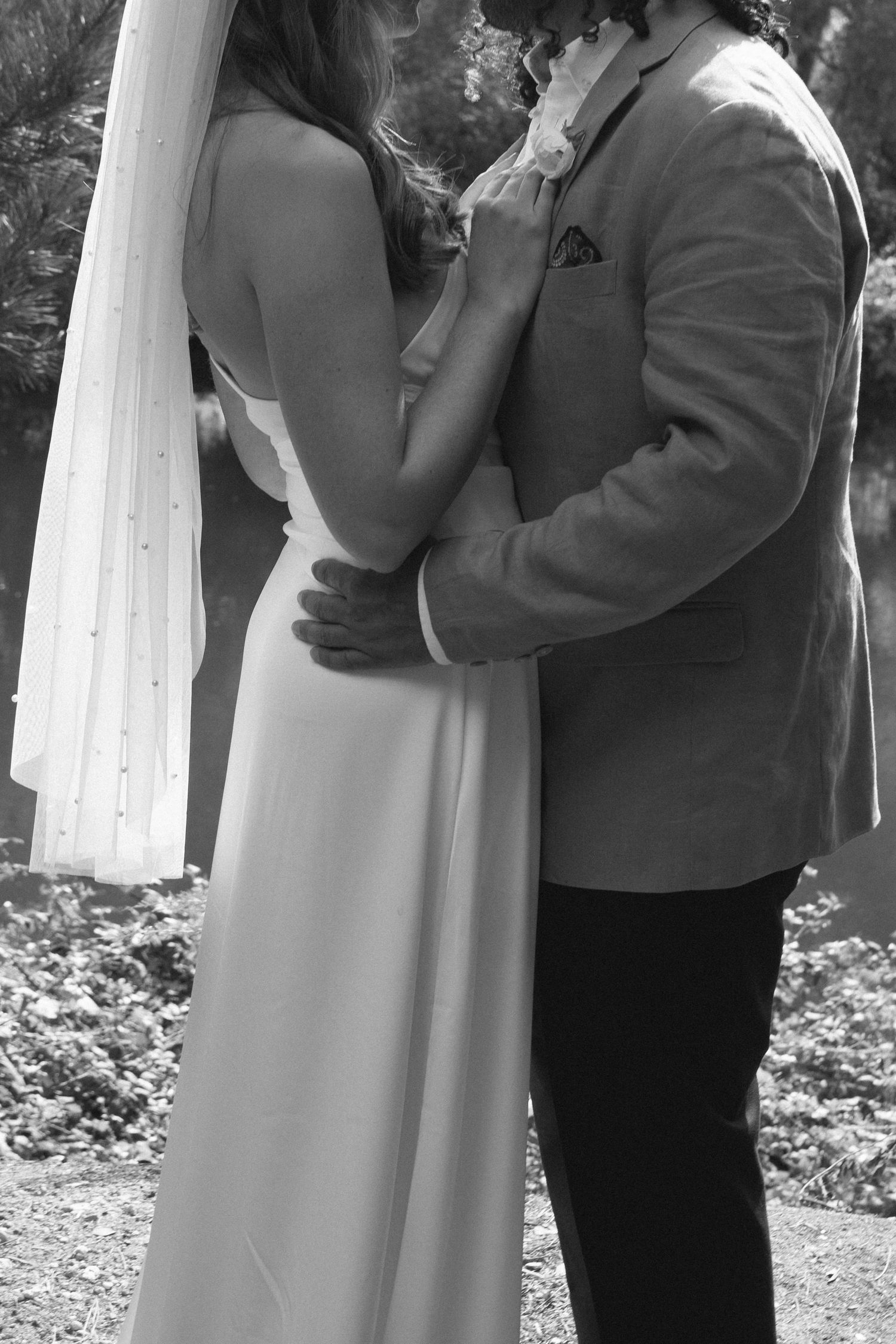 Black and white photo of a bride in white gown and veil embracing a groom in a gray suit outdoors.