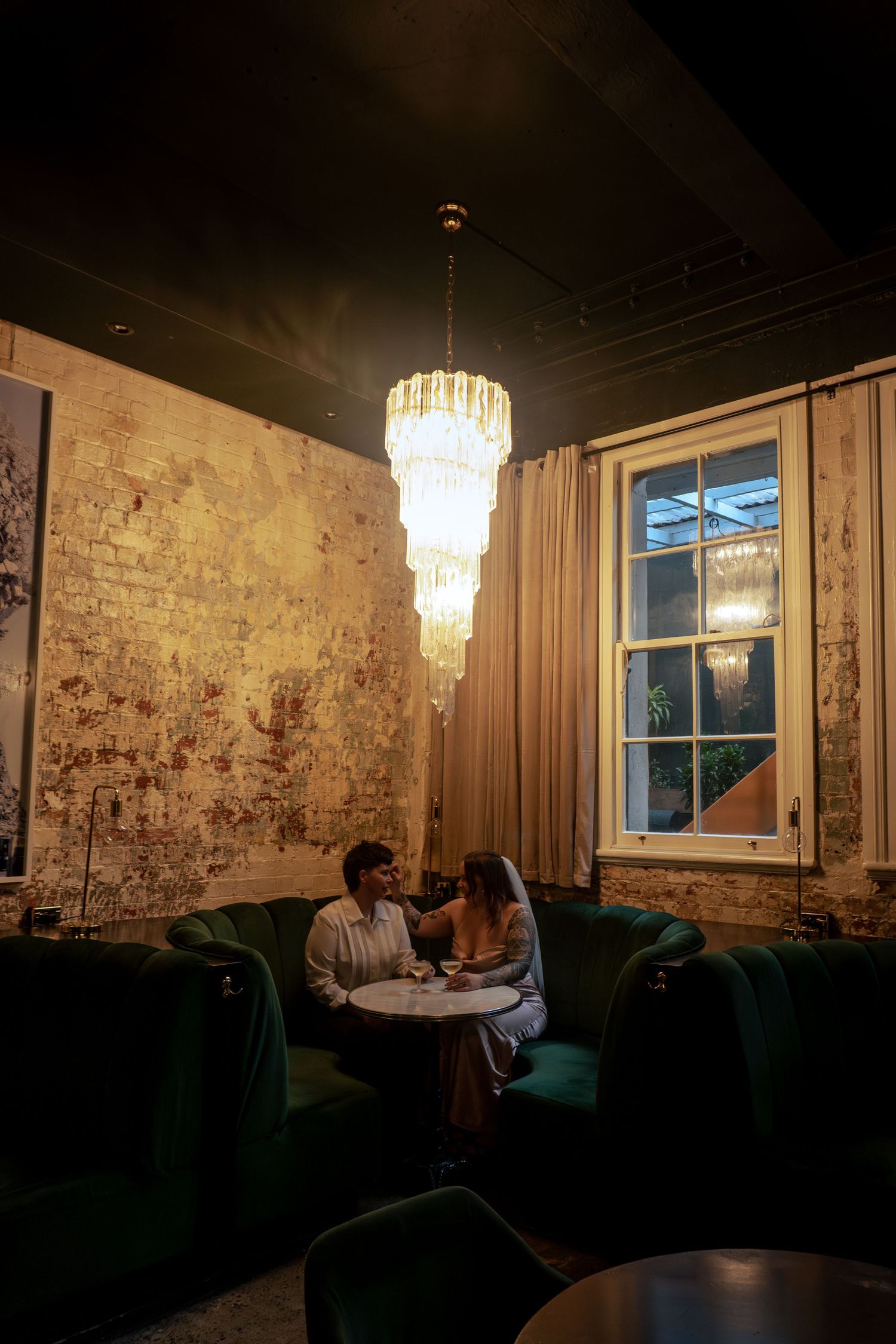 Couple sitting intimately at a marble table beneath a crystal chandelier in a moody, rustic-chic lounge with green velvet chairs.