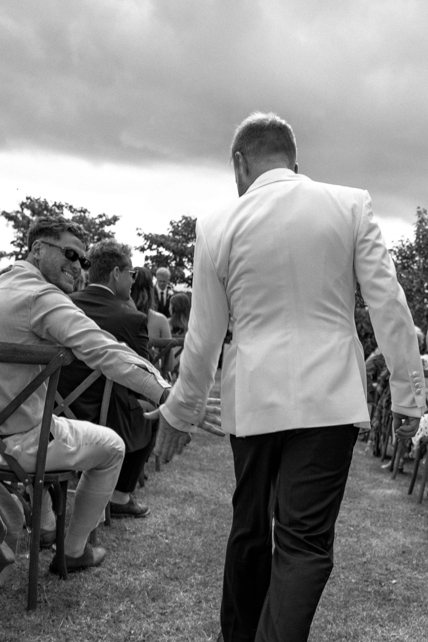 Black and white photo of a man in white jacket walking down wedding aisle past seated guests outdoors.