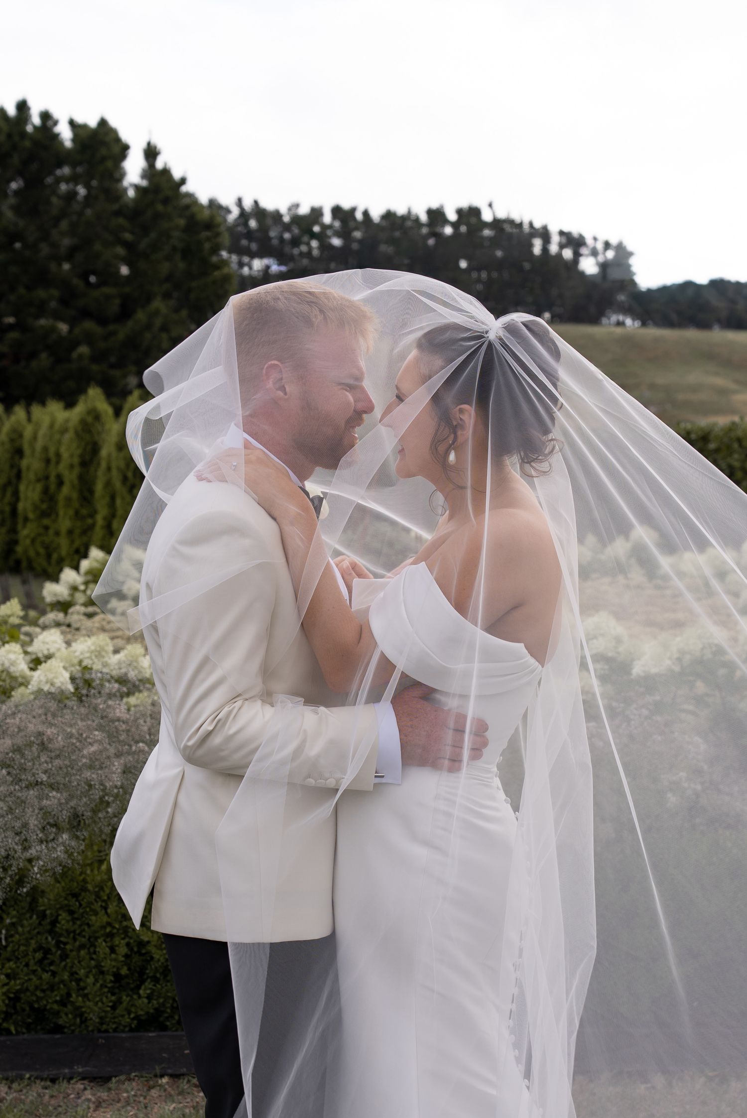 Bride and groom share a romantic moment under a flowing white veil outdoors at their wedding.
