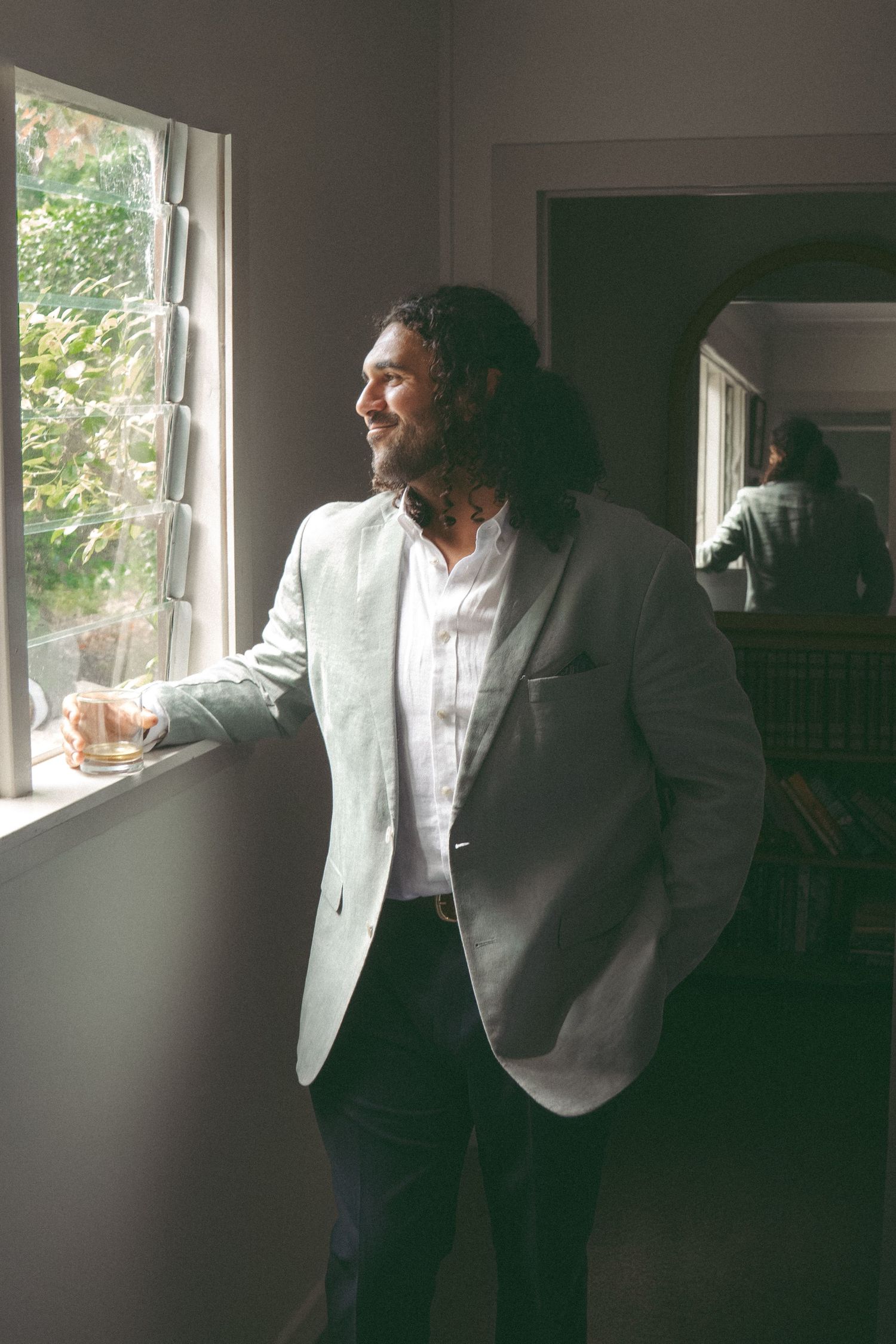 Bearded man with long hair in light blazer gazes out window, holding drink, with mirror reflection visible behind him.