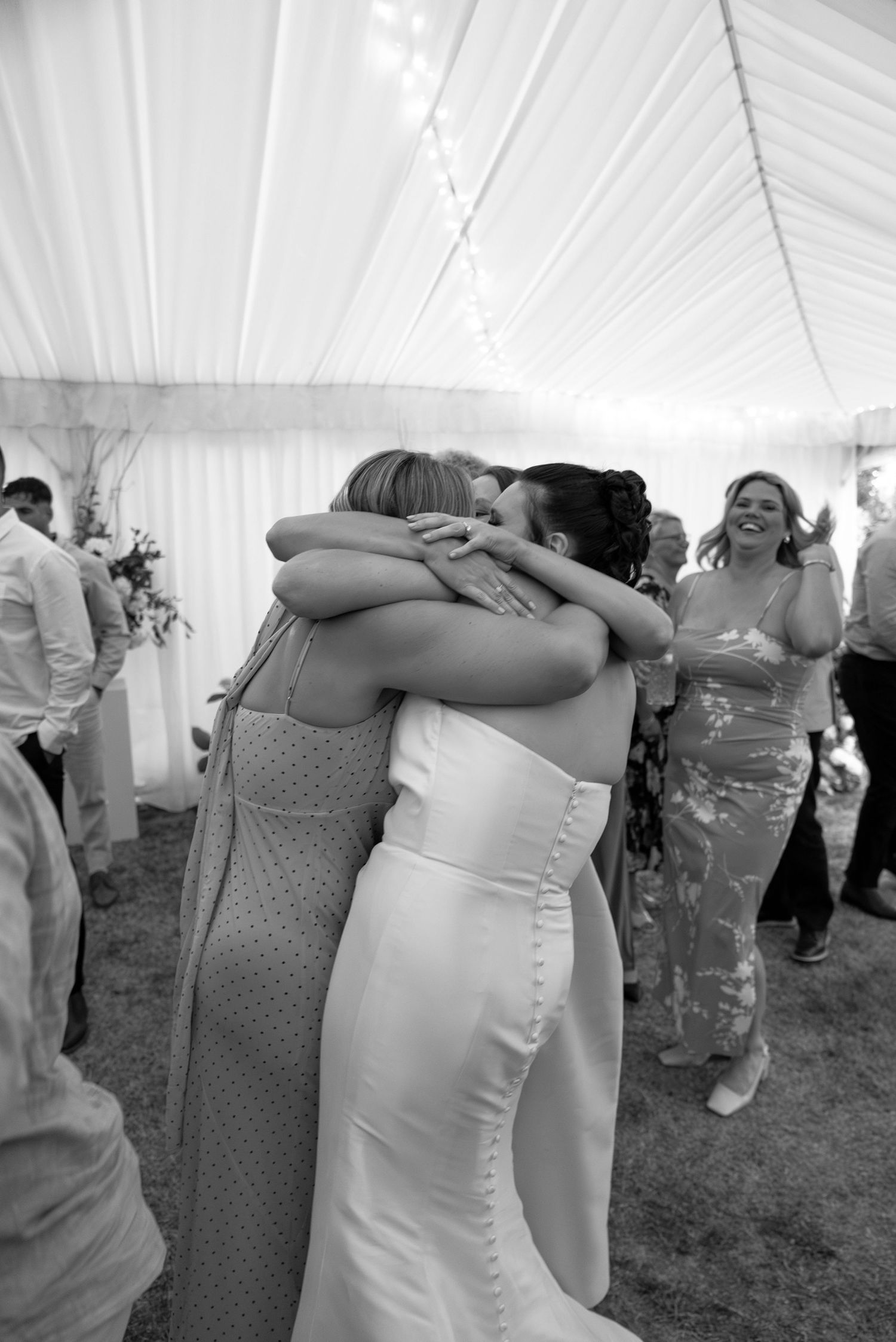 Black and white photo of a bride in a white gown sharing a warm embrace with a guest under a wedding tent.