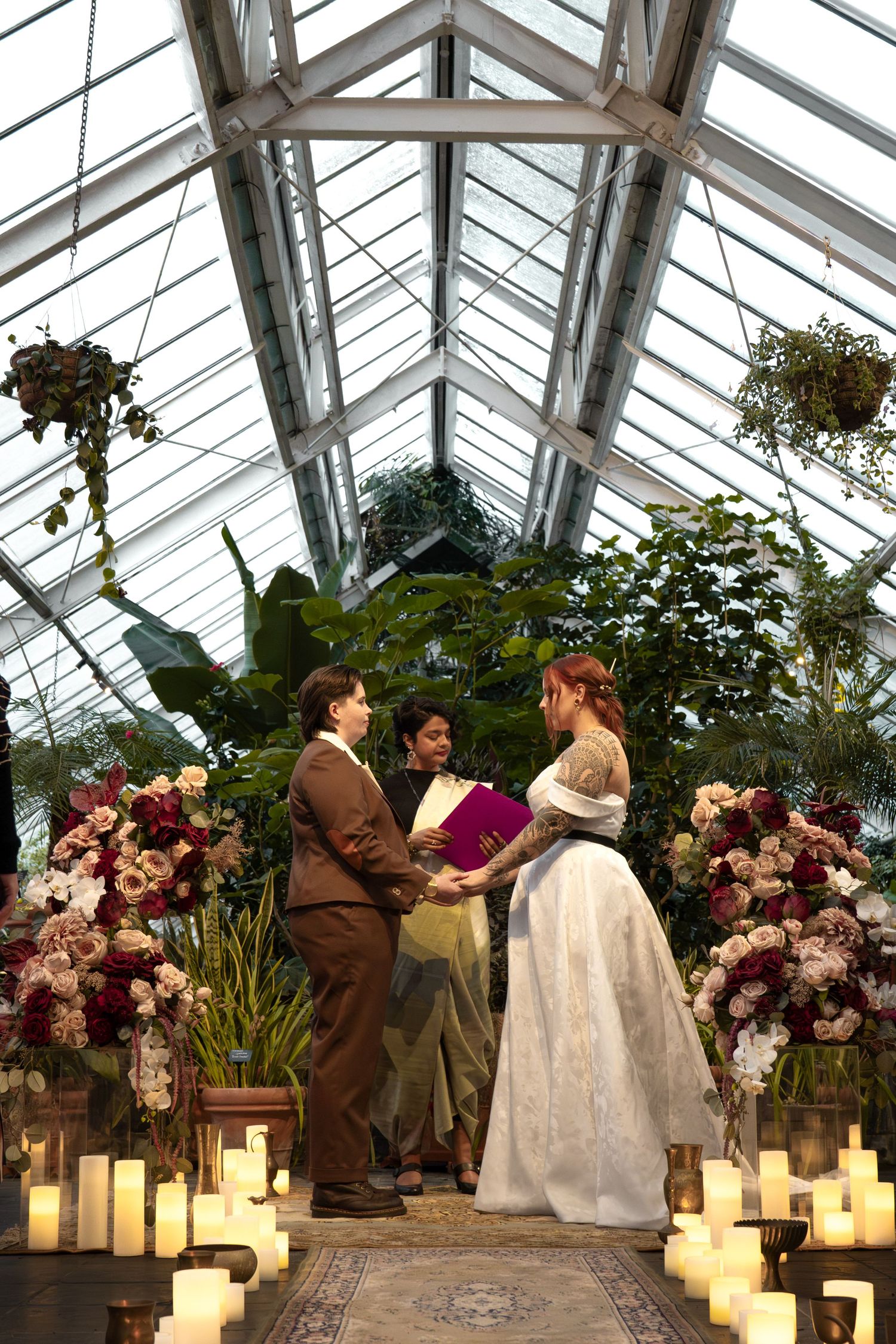 Two brides exchange vows in a candlelit greenhouse wedding ceremony surrounded by lush greenery and romantic floral arrangements.