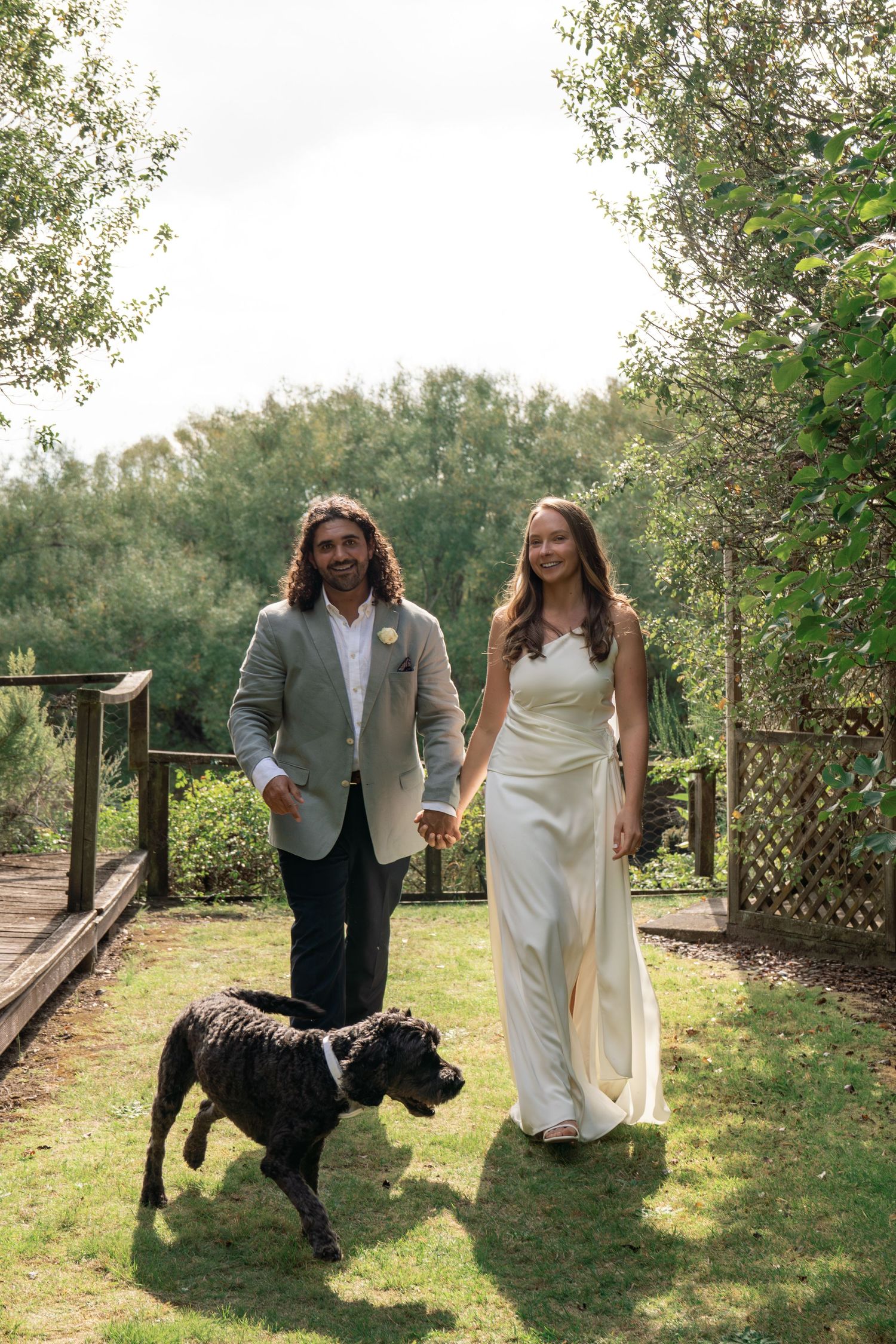 Bride and groom holding hands walking with their dog in a lush garden on their wedding day.