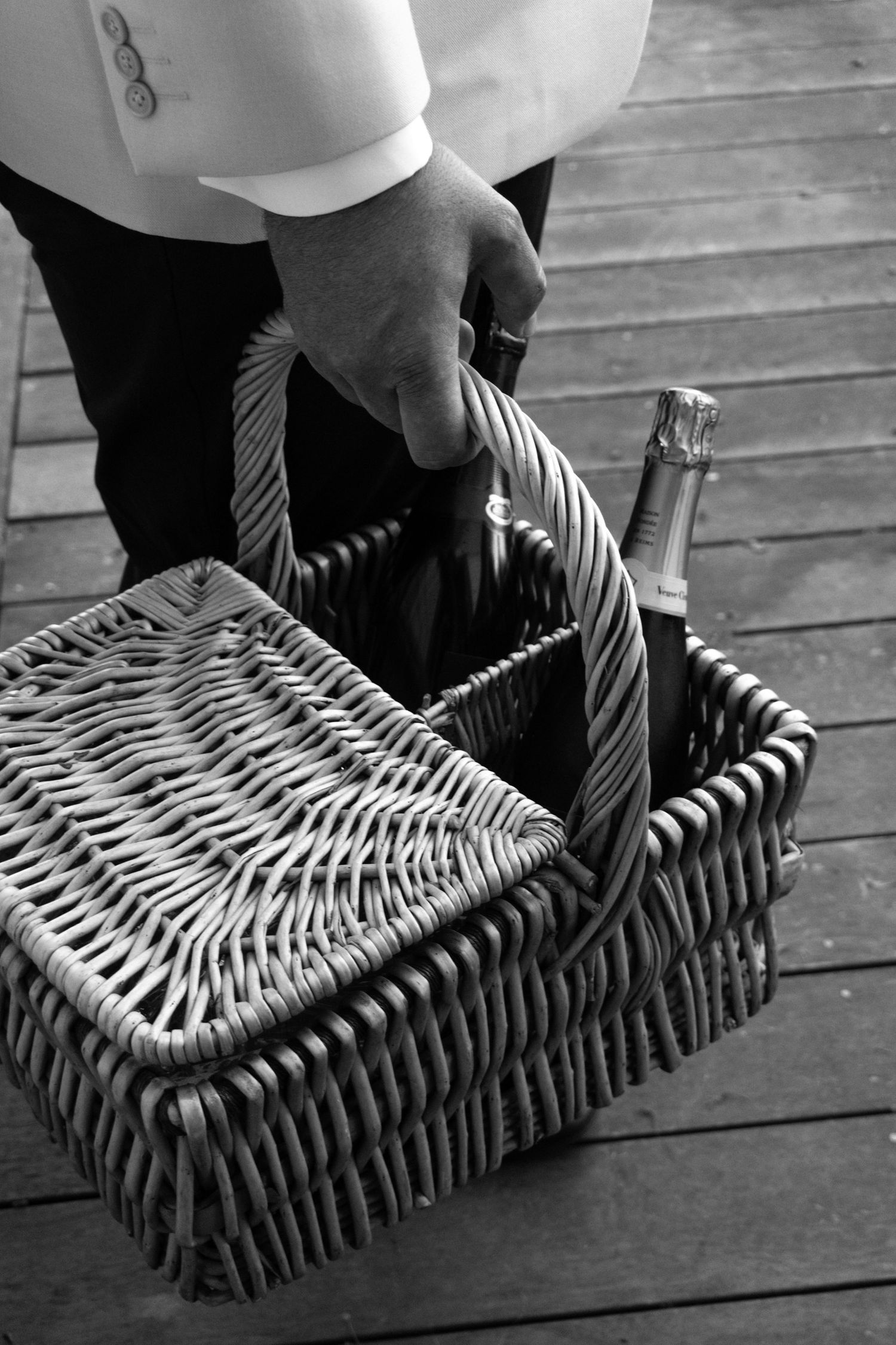 Black and white photo of a man in formal attire carrying a wicker picnic basket with a champagne bottle inside.