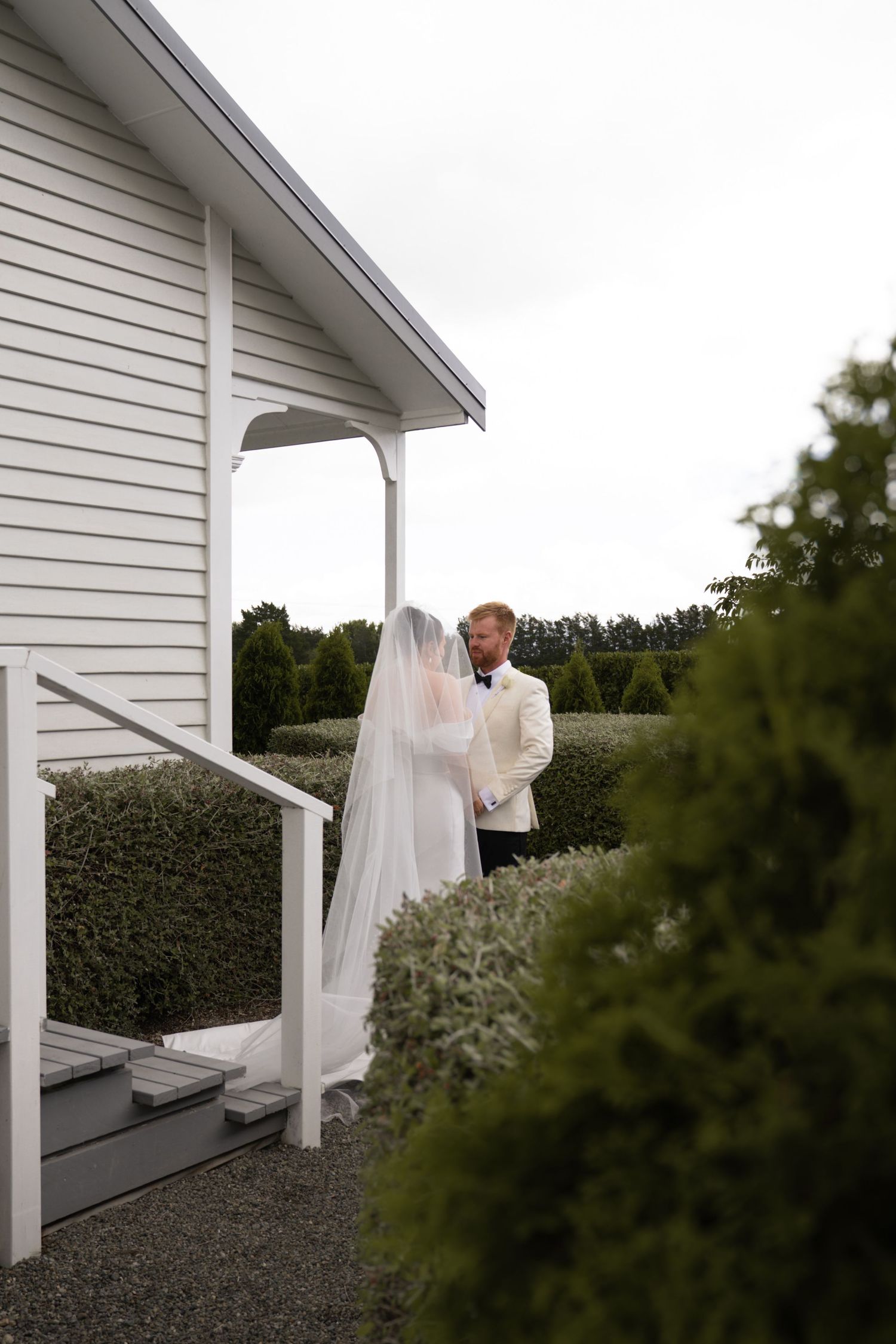 Bride in white gown and veil stands with groom in white jacket near chapel entrance surrounded by manicured hedges.