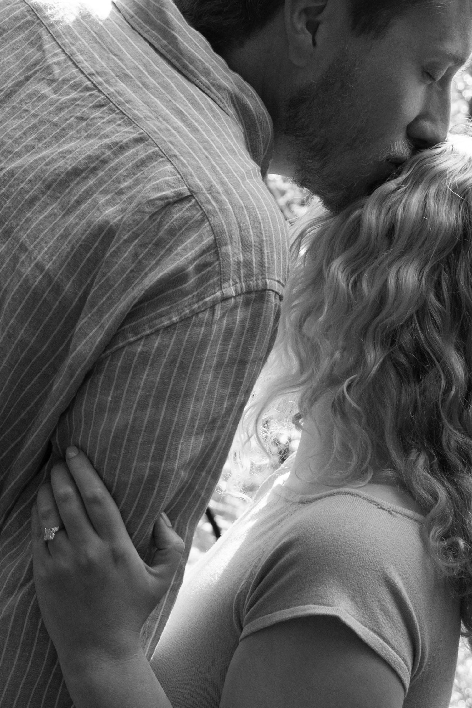 Black and white photo of a couple embracing, man kissing woman's head, her hand showing an engagement ring.